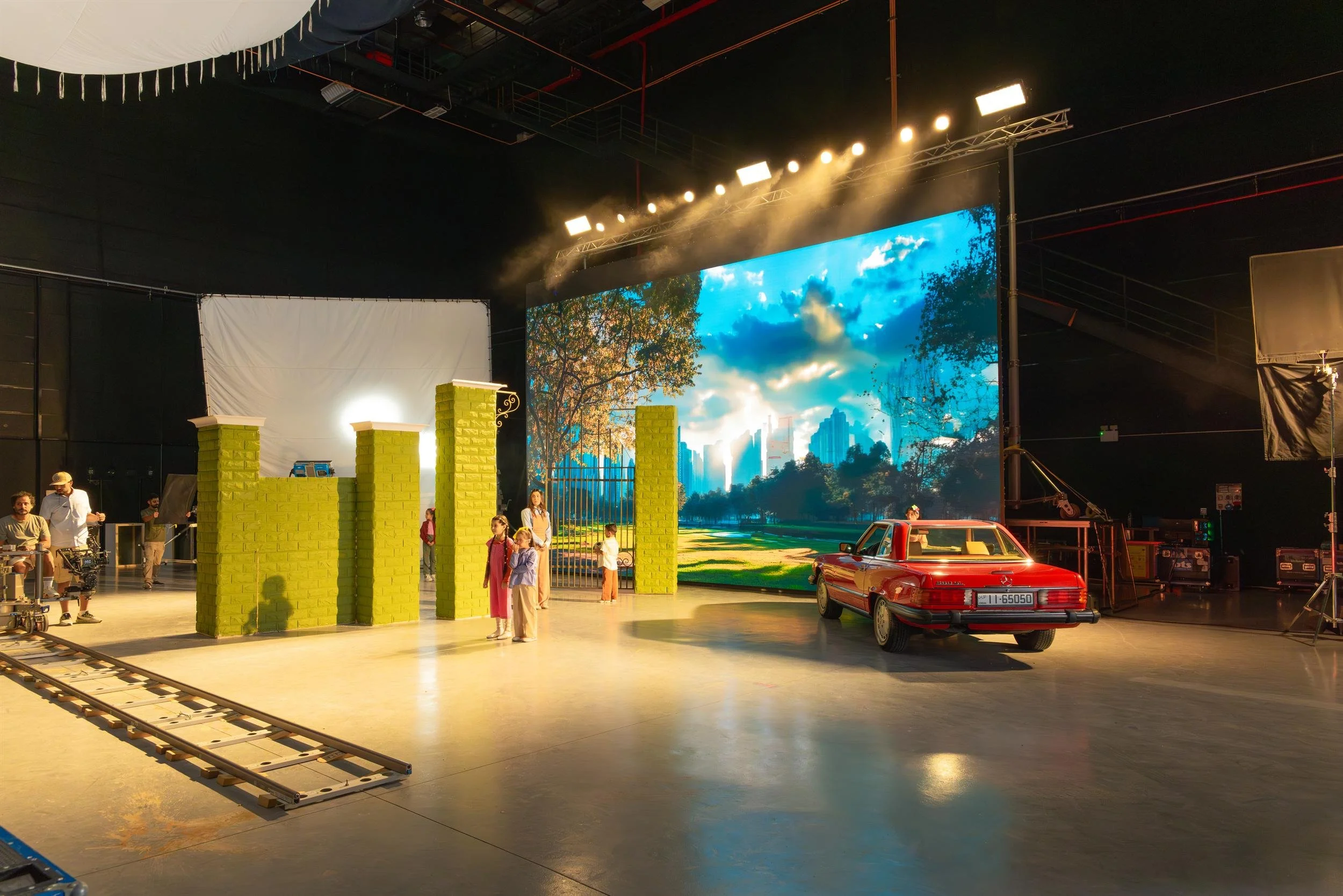 Ahlan Simsim unit still: A studio set with children and a red car in front of a large LED screen showing a city park.