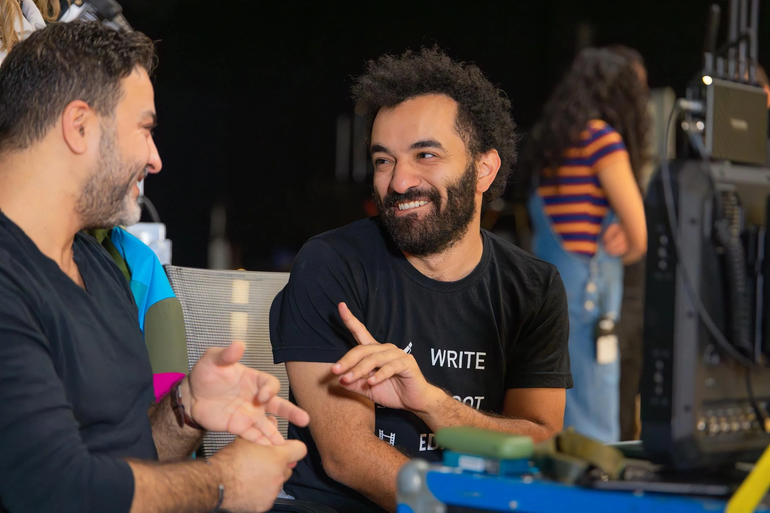Ahlan Simsim unit still: Two crew members smile and talk on a dark film set.
