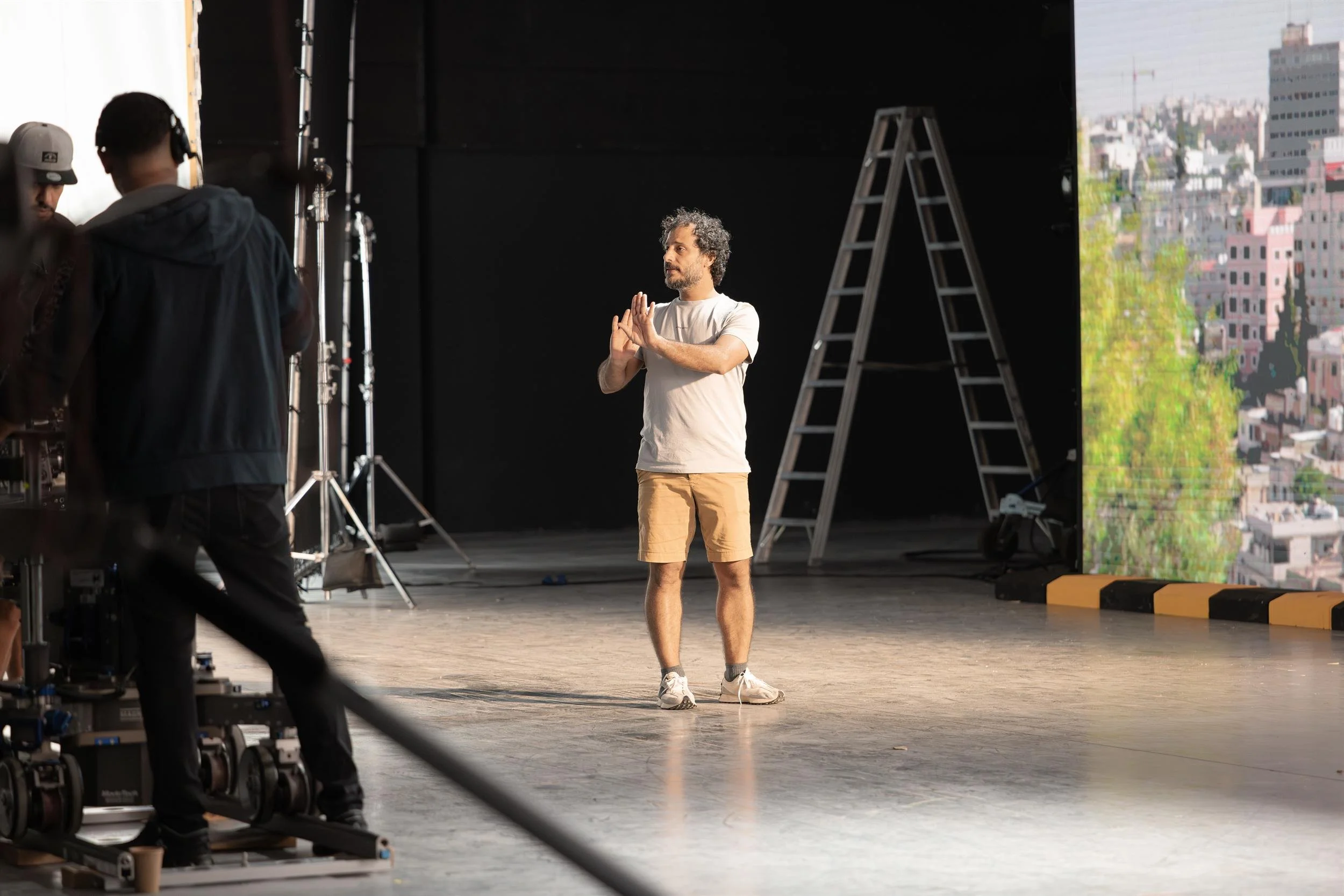 Ahlan Simsim unit still: a man gestures on set in front of an LED cityscape and a silver ladder.
