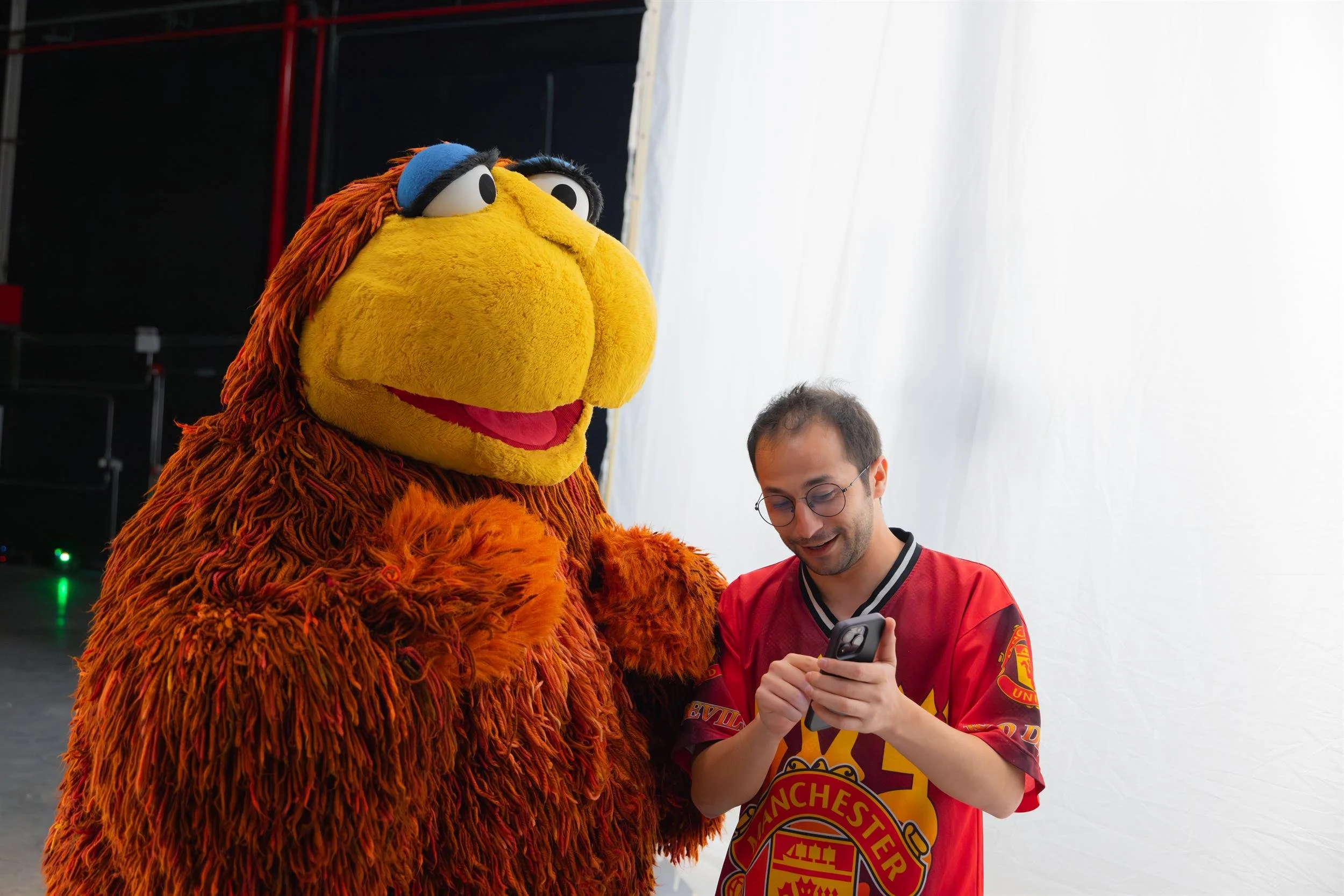 Ahlan Simsim unit still: a man in a red jersey looks at his phone beside a large brown Muppet.