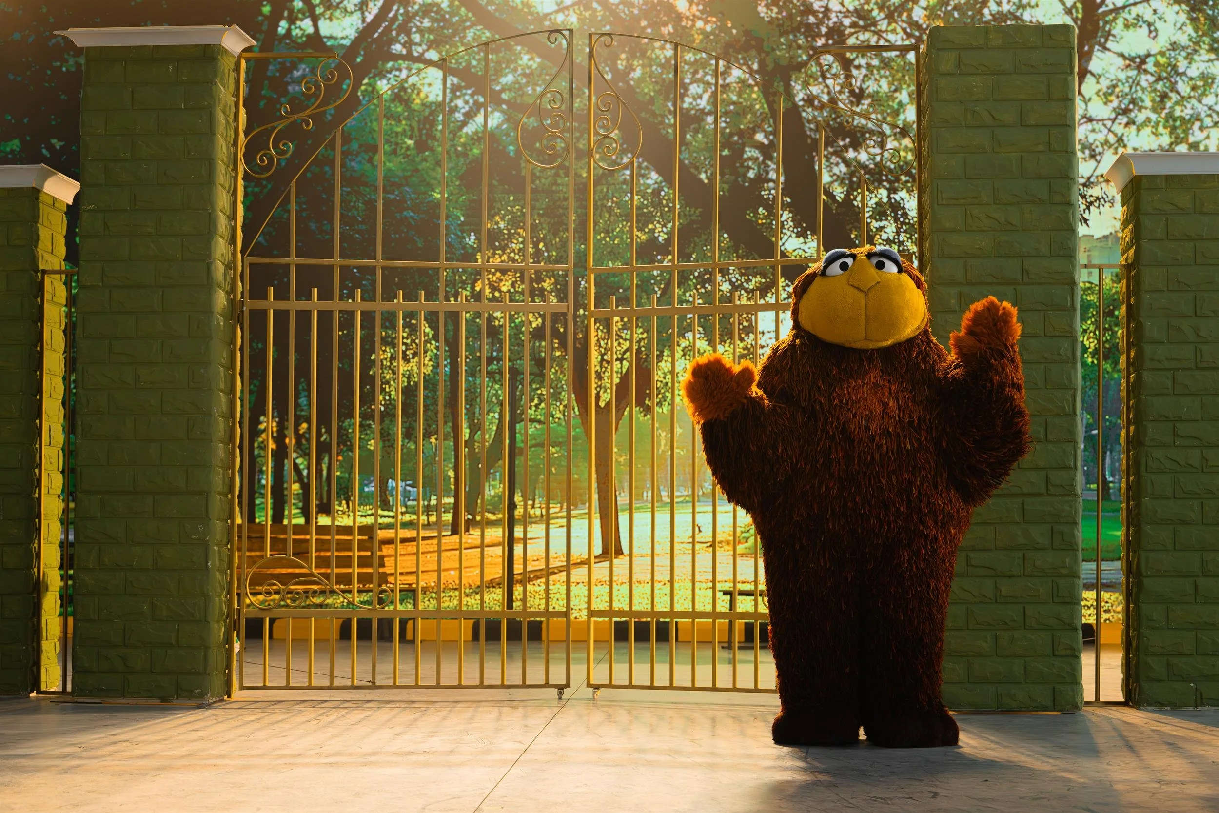 Ahlan Simsim key art: the brown muppet waves at the park gate as children run past in the sunlight.