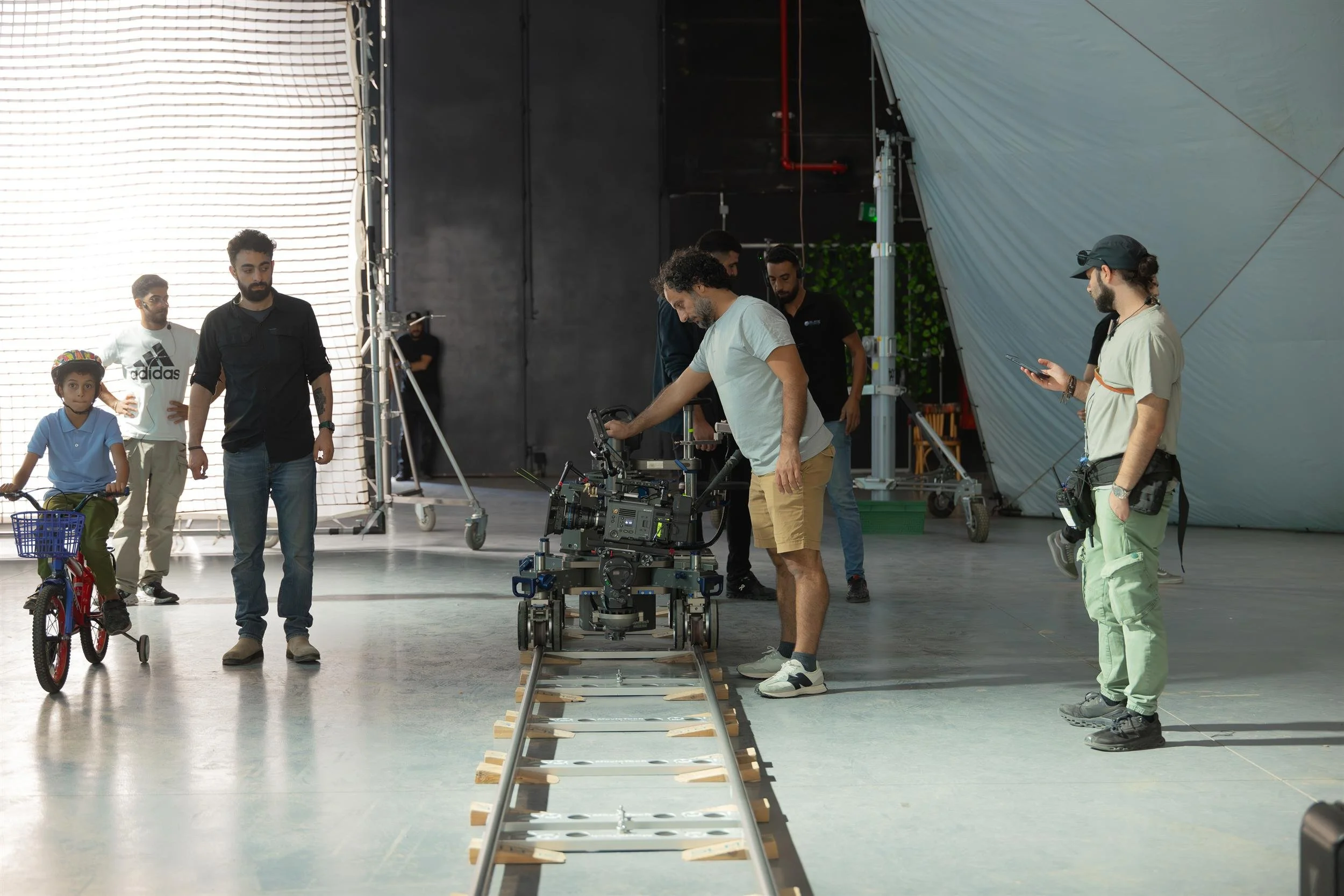 Ahlan Simsim unit still: a crew member adjusts a camera on tracks while a boy on a red bicycle waits nearby.