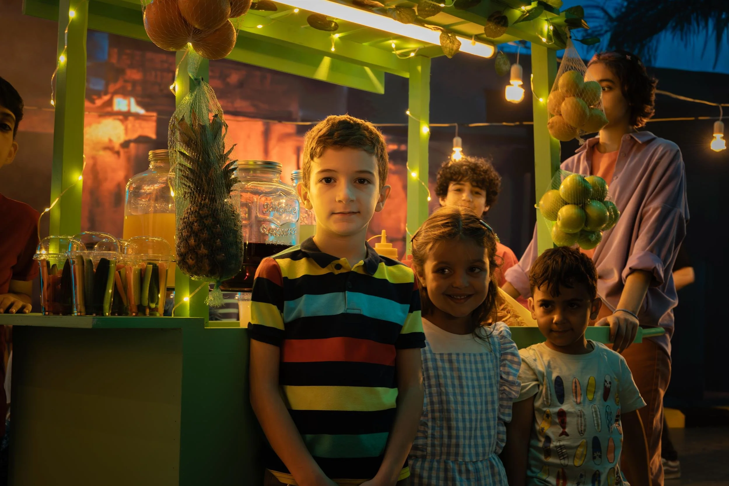 Ahlan Simsim unit still: three children stand in front of a brightly lit fruit-juice stall at night.