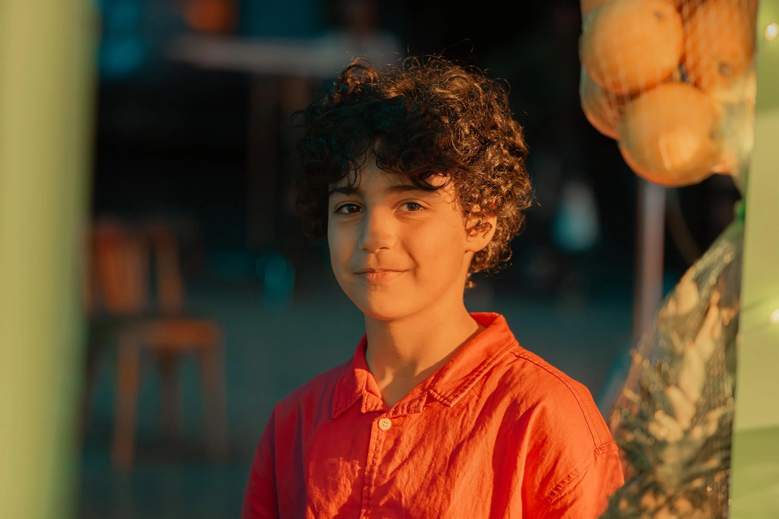 Ahlan Simsim unit still: a smiling boy in a red shirt at a night market with a bag of oranges nearby.