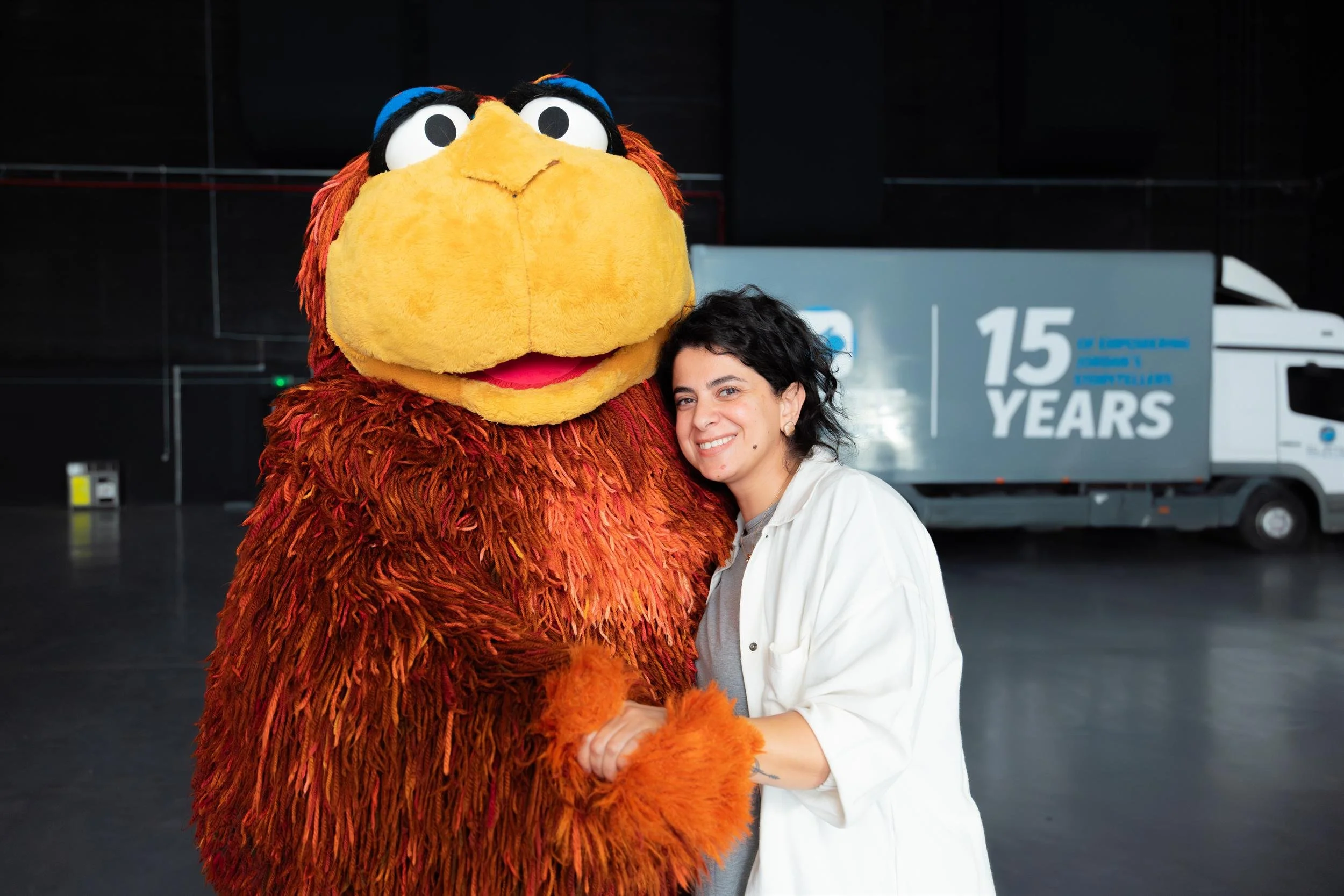 Ahlan Simsim unit still: a woman smiles while hugging a large shaggy brown Muppet in a studio.