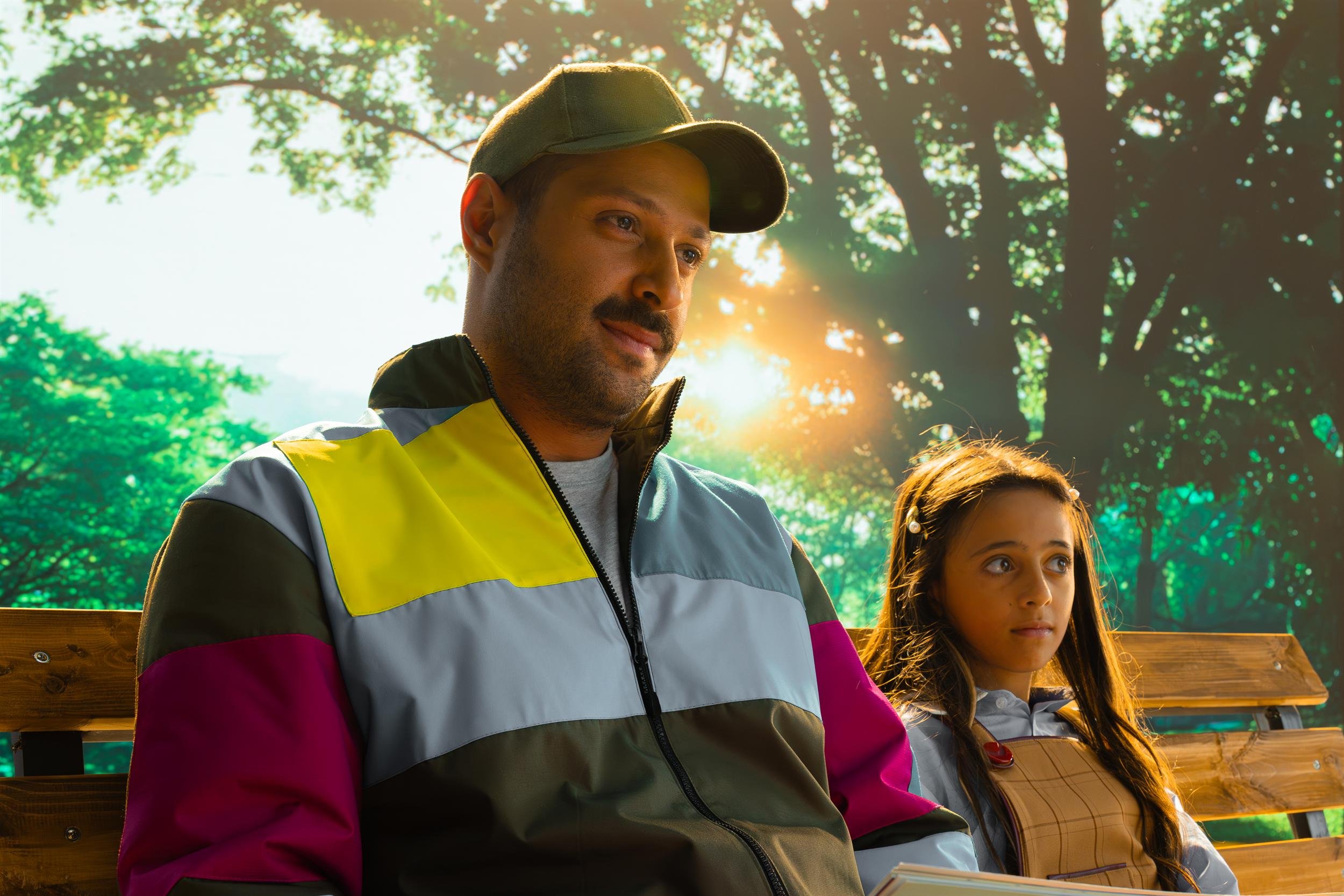Ahlan Simsim unit still: A man in a colorful jacket and a young girl sit side by side on a wooden park bench.