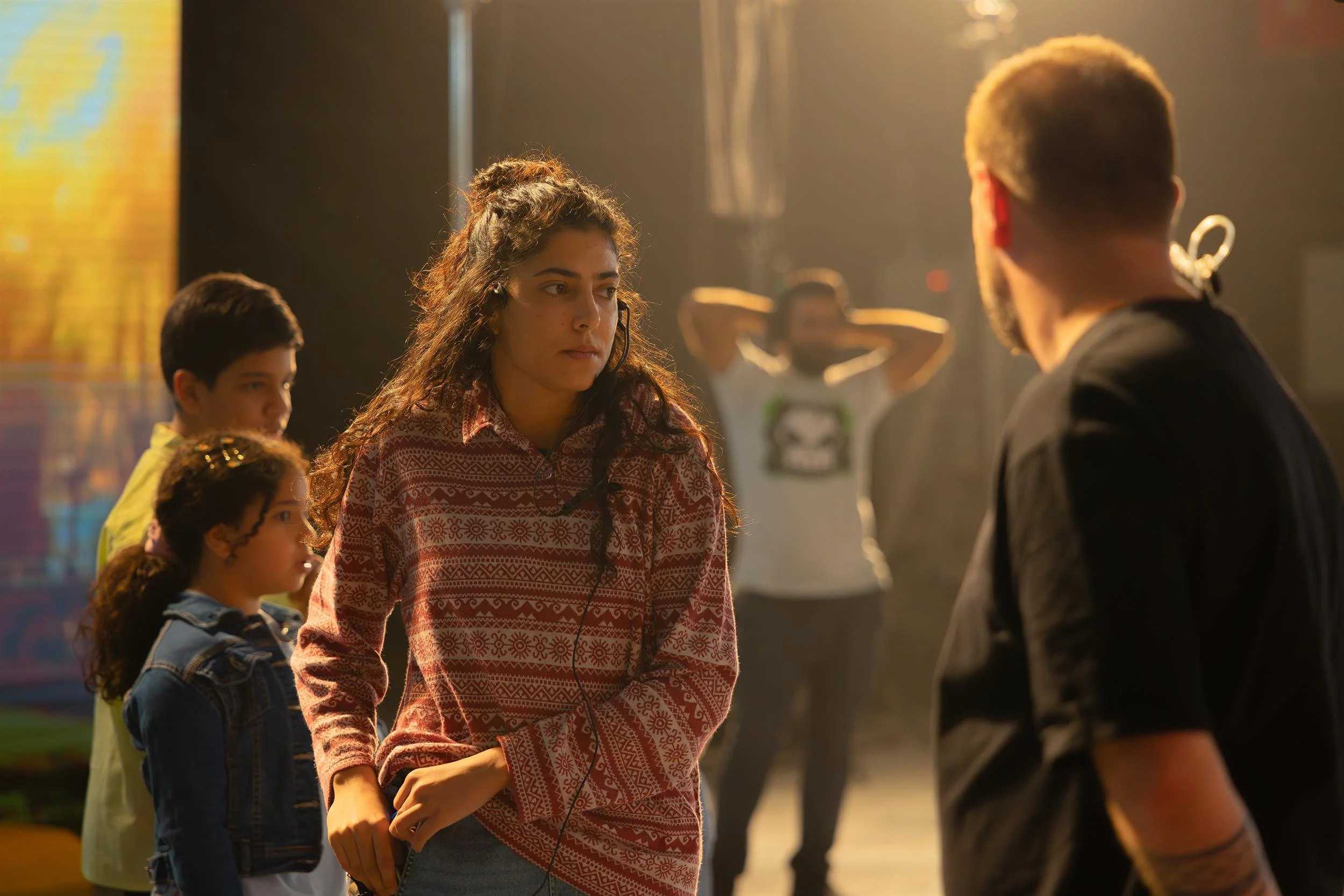 Ahlan Simsim unit still: a woman in a red sweater and headset talks on set beside two children.