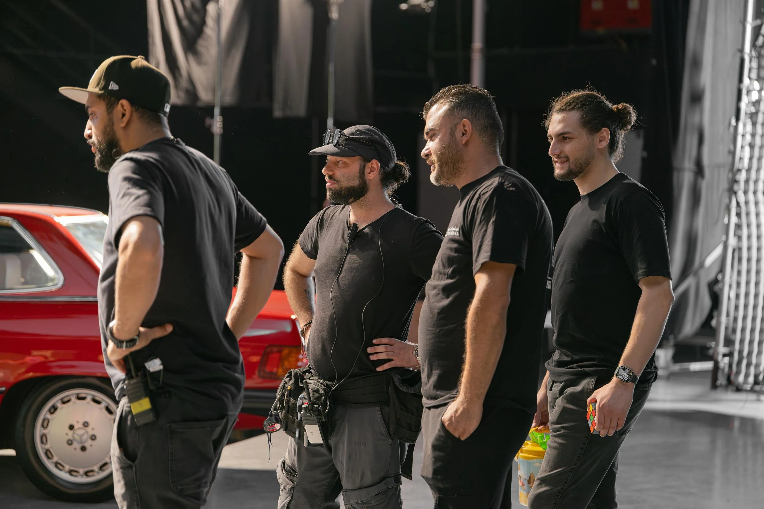 Ahlan Simsim unit still: Four crew members in black shirts stand together on a studio set beside a vintage red car.