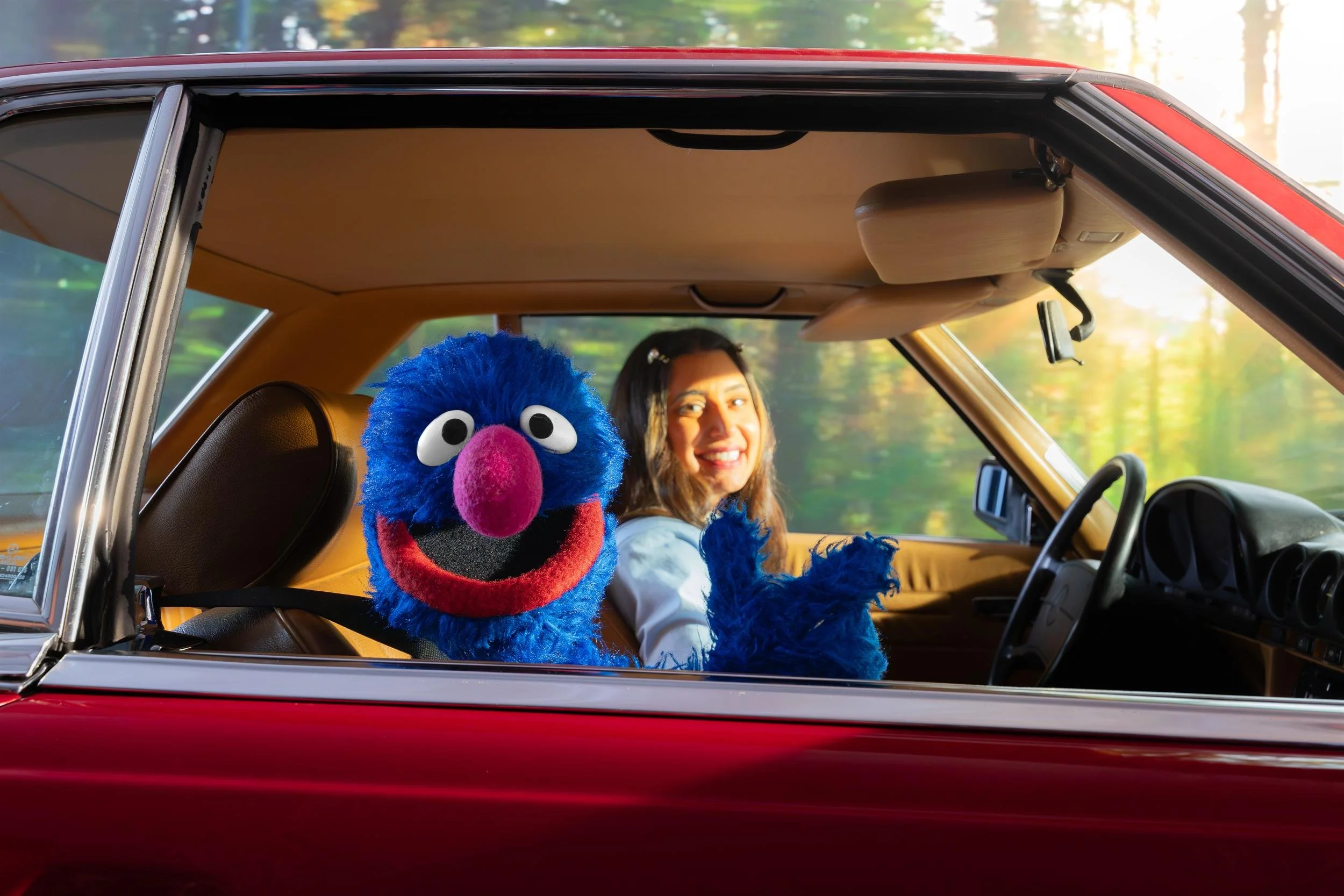 Ahlan Simsim key art: A woman and a blue Muppet smile from inside a vintage red car.