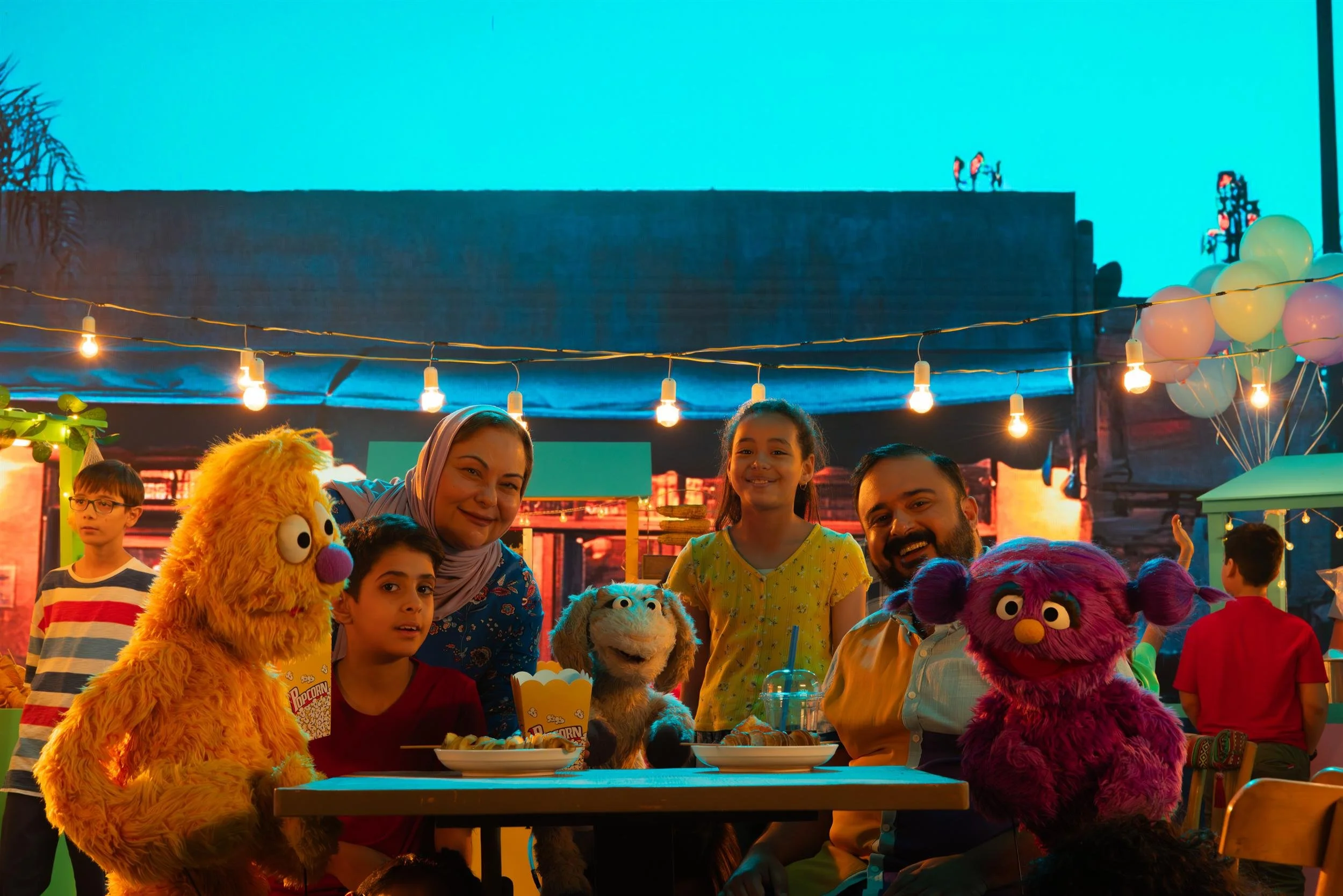 Ahlan Simsim unit still: three Muppets sit with children and adults at a night-market table under string lights.