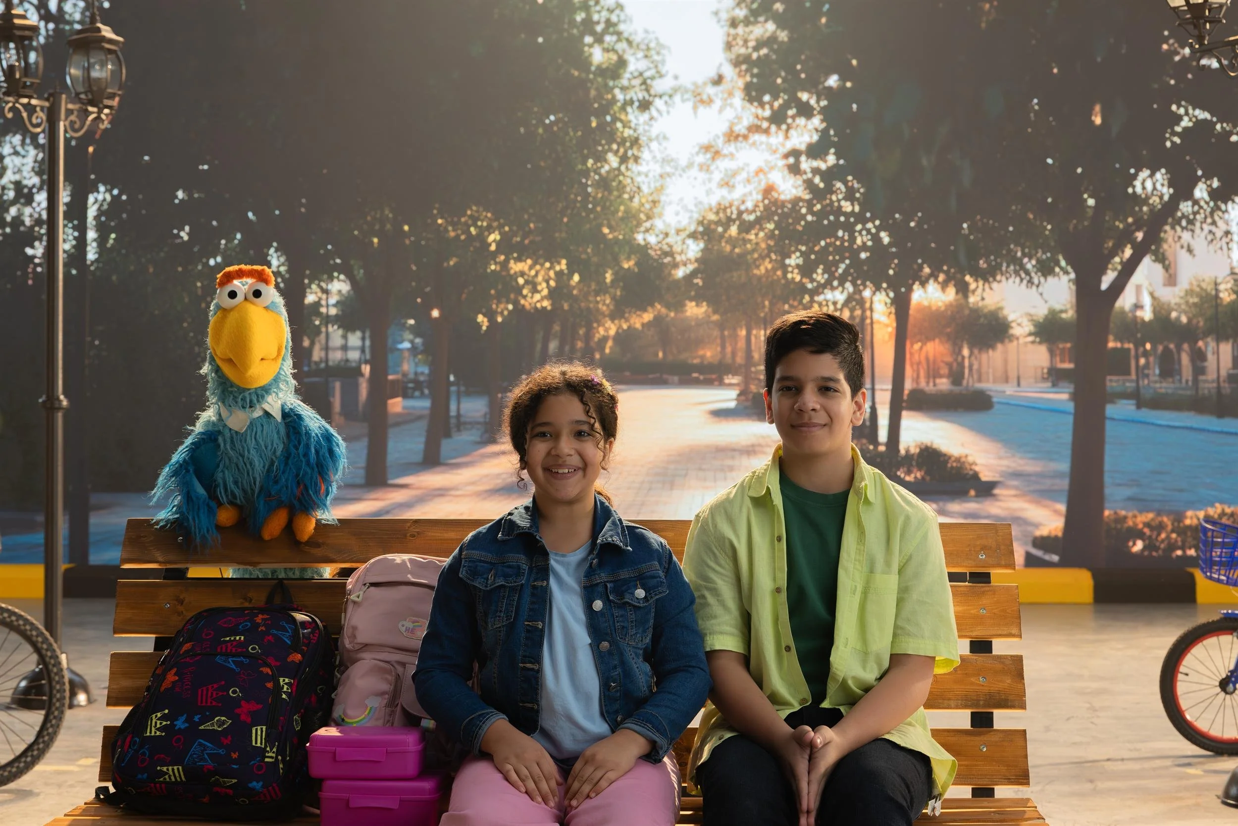 Ahlan Simsim unit still: Two children and a blue bird Muppet sit on a park bench with school backpacks.