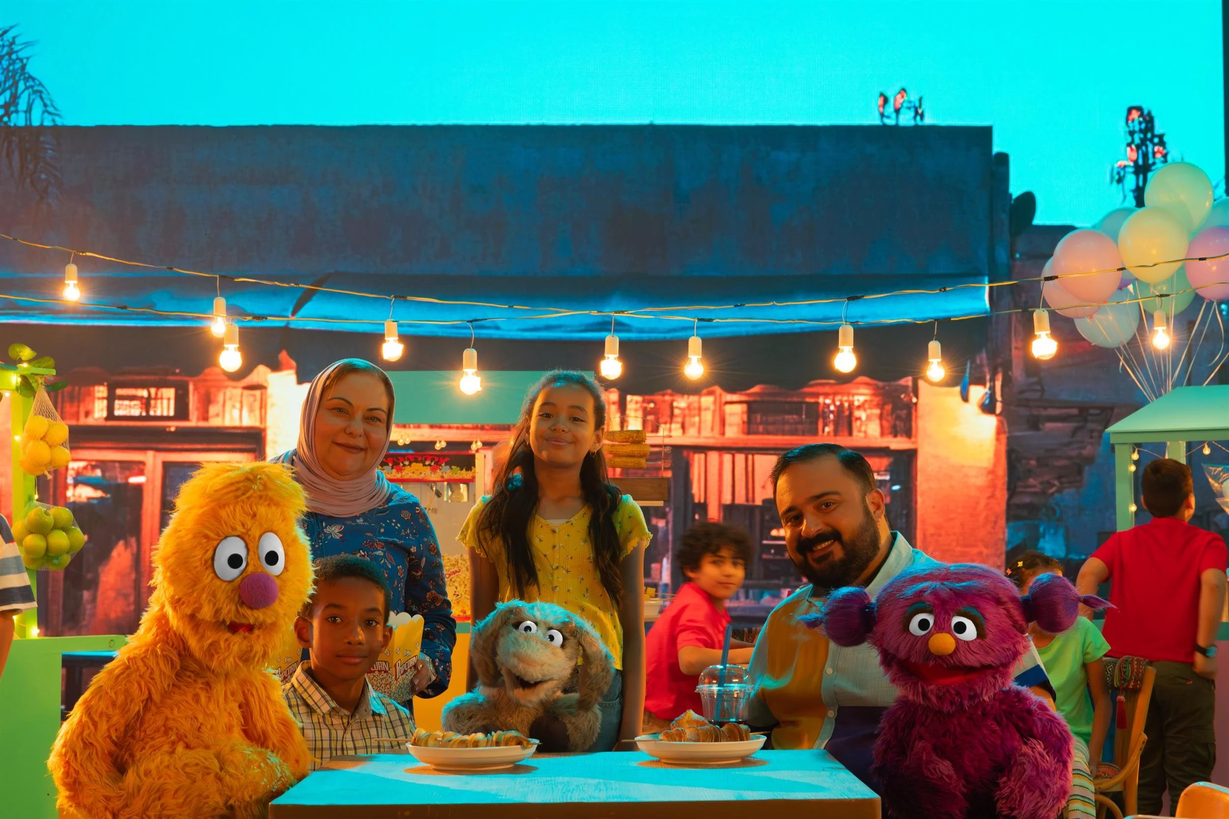 Ahlan Simsim key art: Cast members and orange, grey, and purple Muppets pose together at a nighttime street festival.
