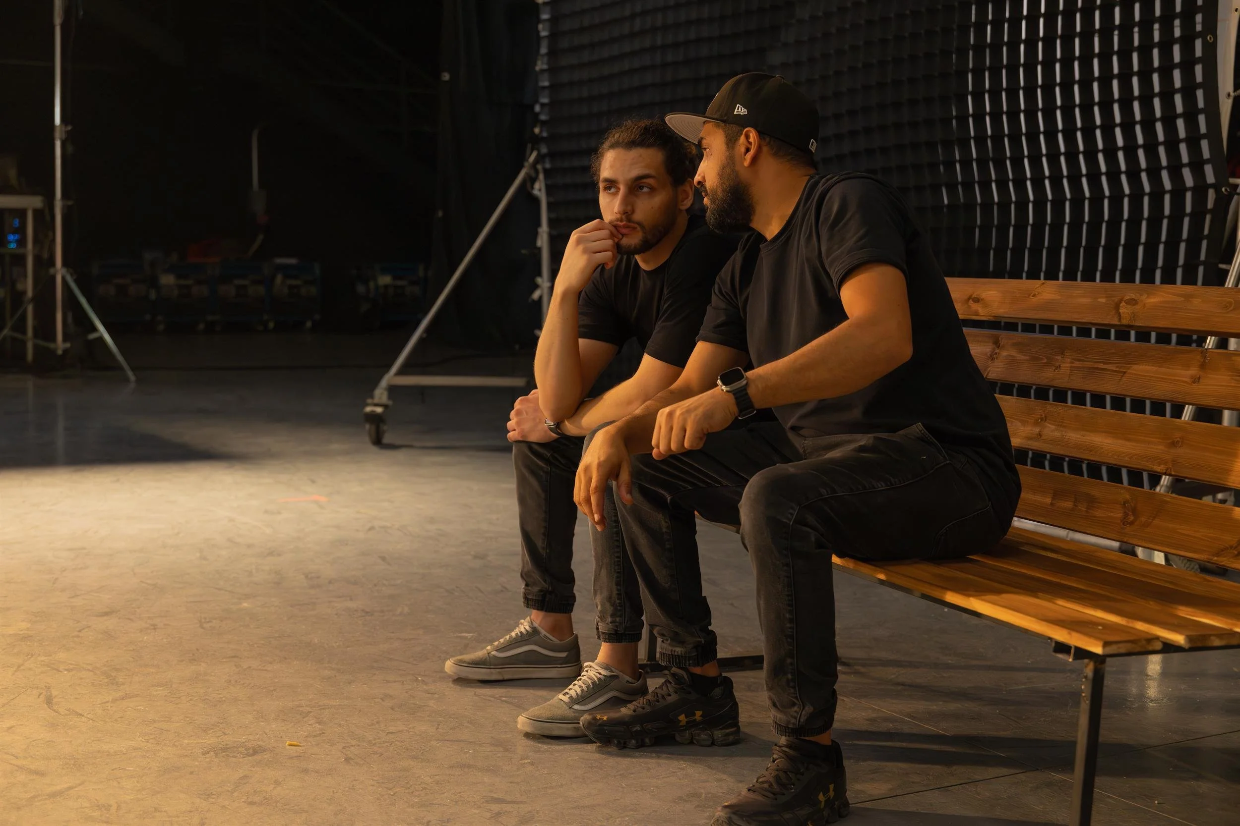 Ahlan Simsim unit still: two crew members in black sit on a wooden bench in a dim studio.