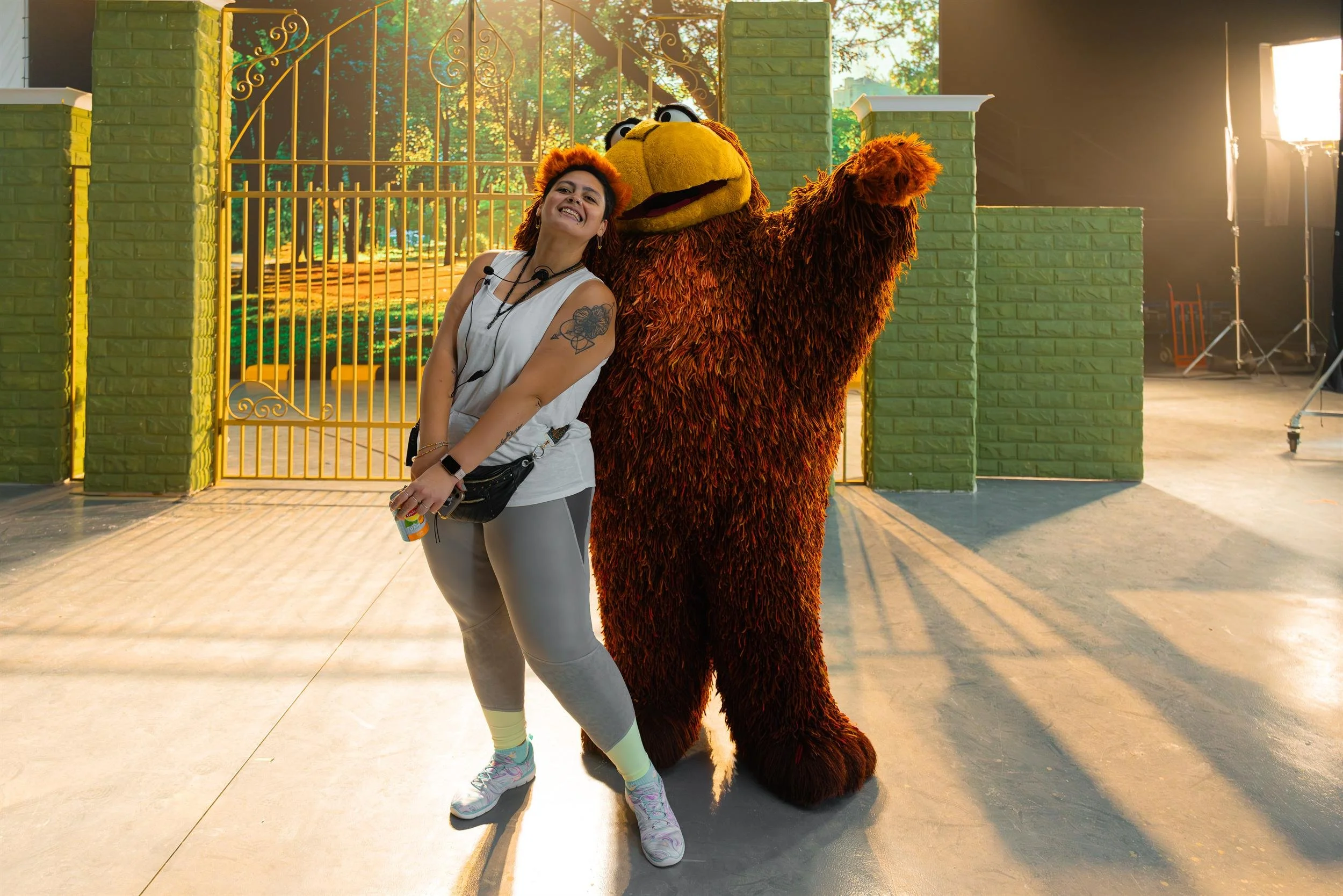 Ahlan Simsim unit still: A smiling crew member poses with a large brown Muppet waving on a park-gate studio set.