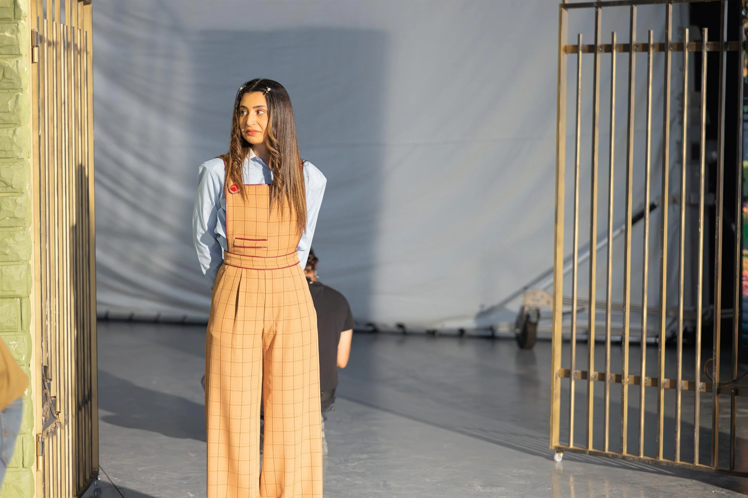 Ahlan Simsim unit still: A woman in tan overalls stands between golden metal gates on a studio set.