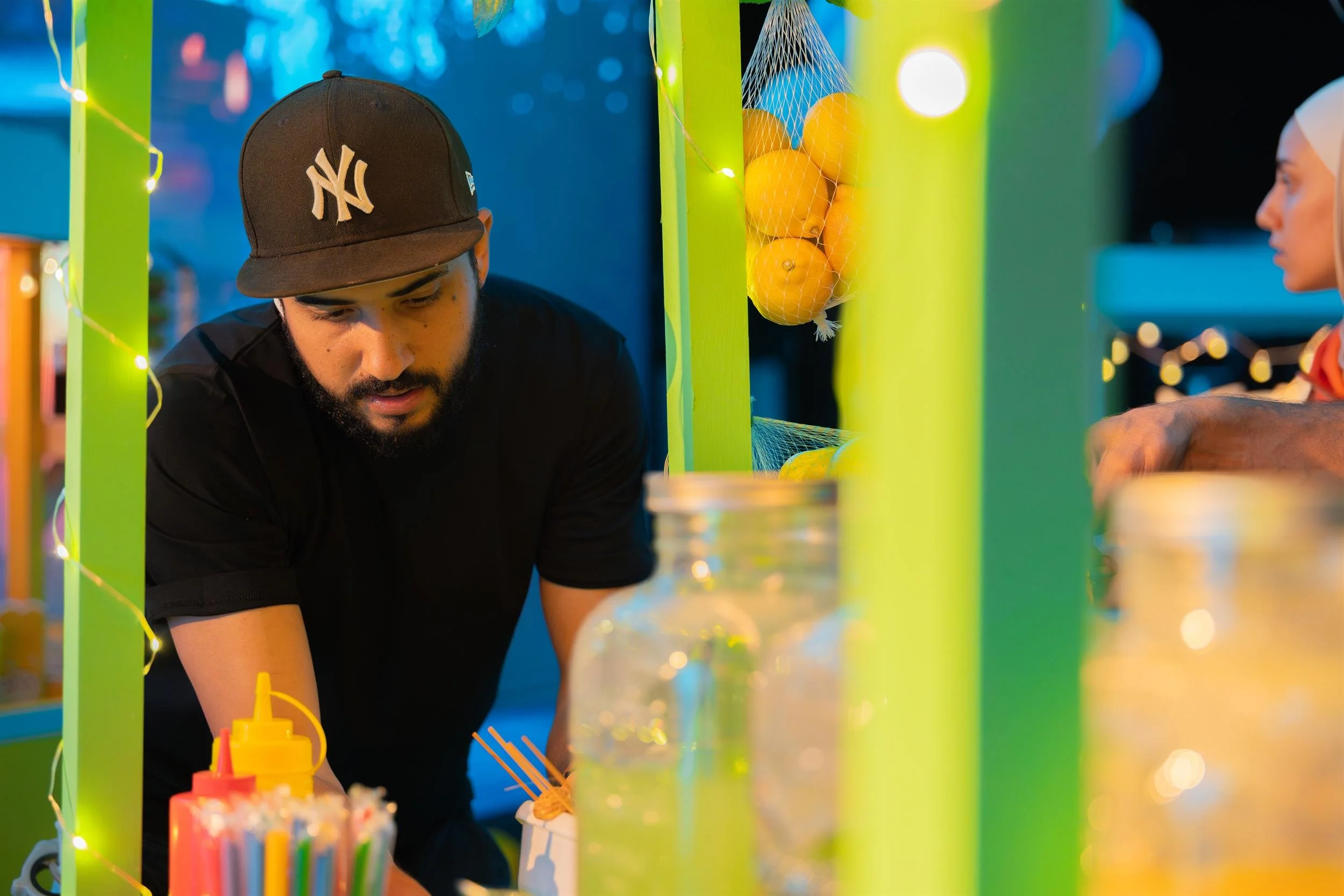 Ahlan Simsim unit still: a man in a baseball cap works behind a green market stall with string lights and lemons.