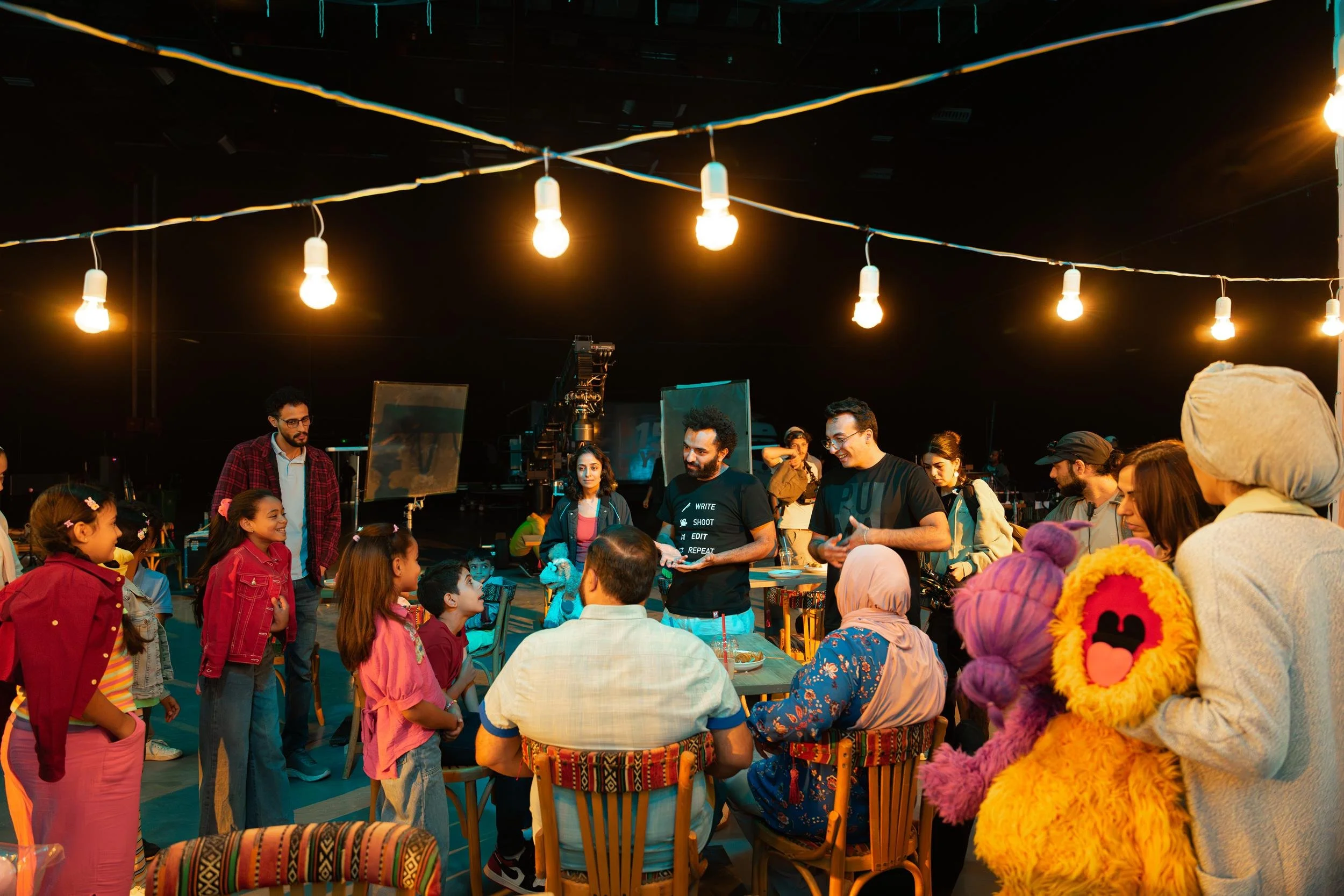 Ahlan Simsim unit still: crew, children, and Muppets gather under string lights on a night-market set.
