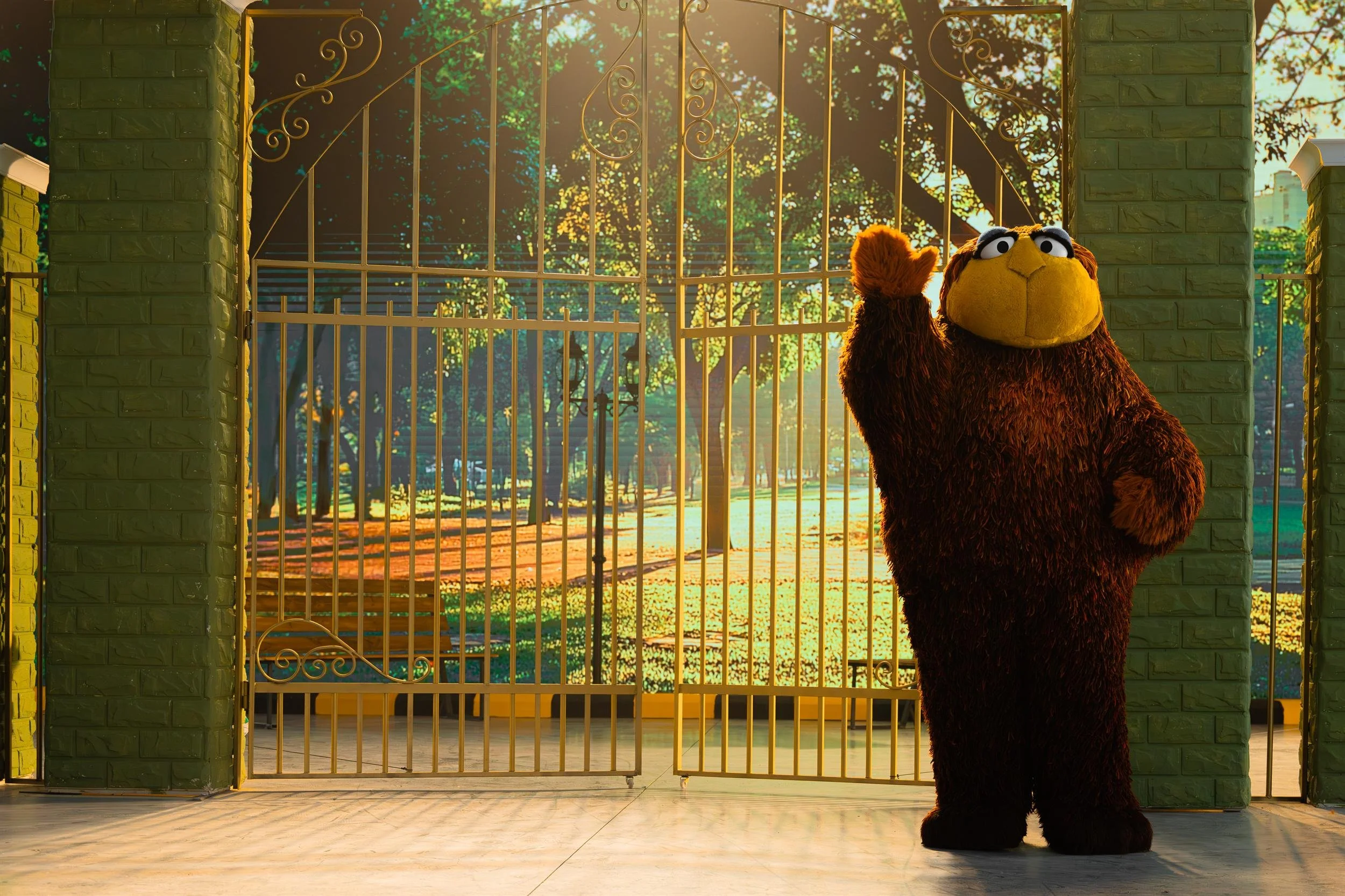 Ahlan Simsim key art: a large brown muppet stands at the wrought-iron park gate on the Amman set.