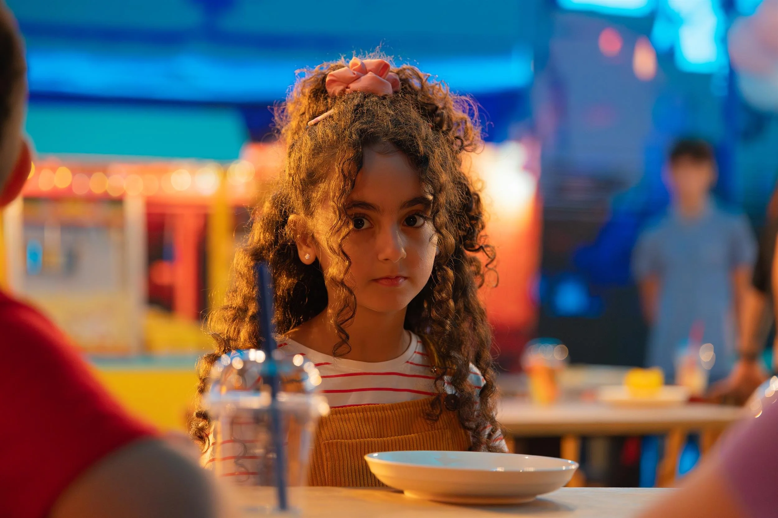 Ahlan Simsim unit still: a girl in yellow overalls sits at a cafe table on a colorful night set.