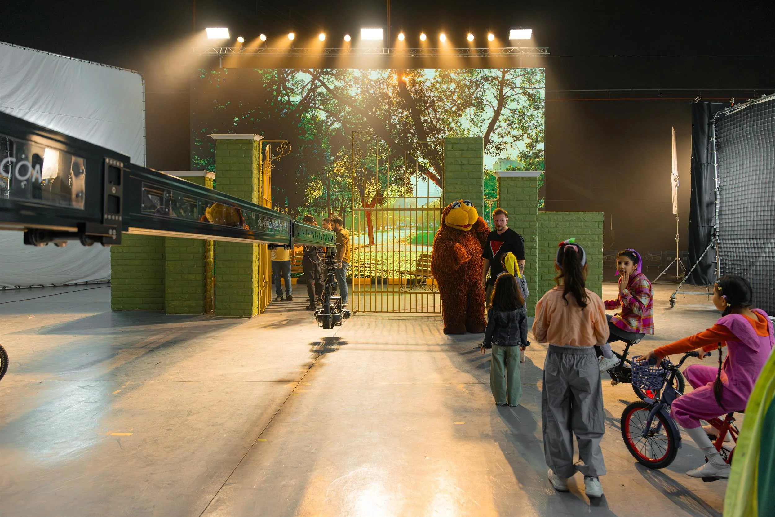 Ahlan Simsim unit still: A wide studio view of a park-gate set, a brown Muppet, a camera crane, and children near a bicycle.