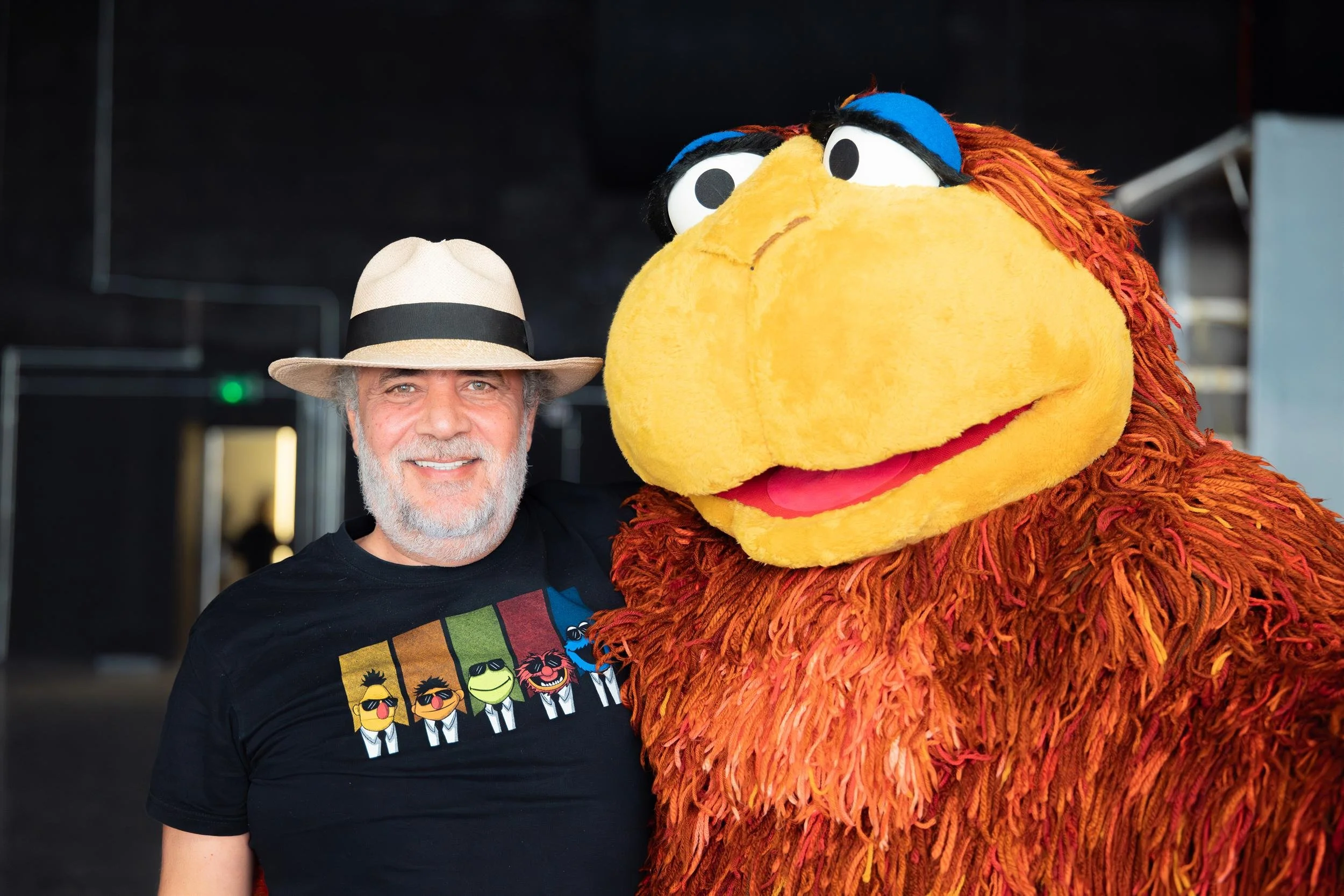 Ahlan Simsim unit still: a man in a fedora and Muppet t-shirt stands next to a large shaggy brown Muppet.