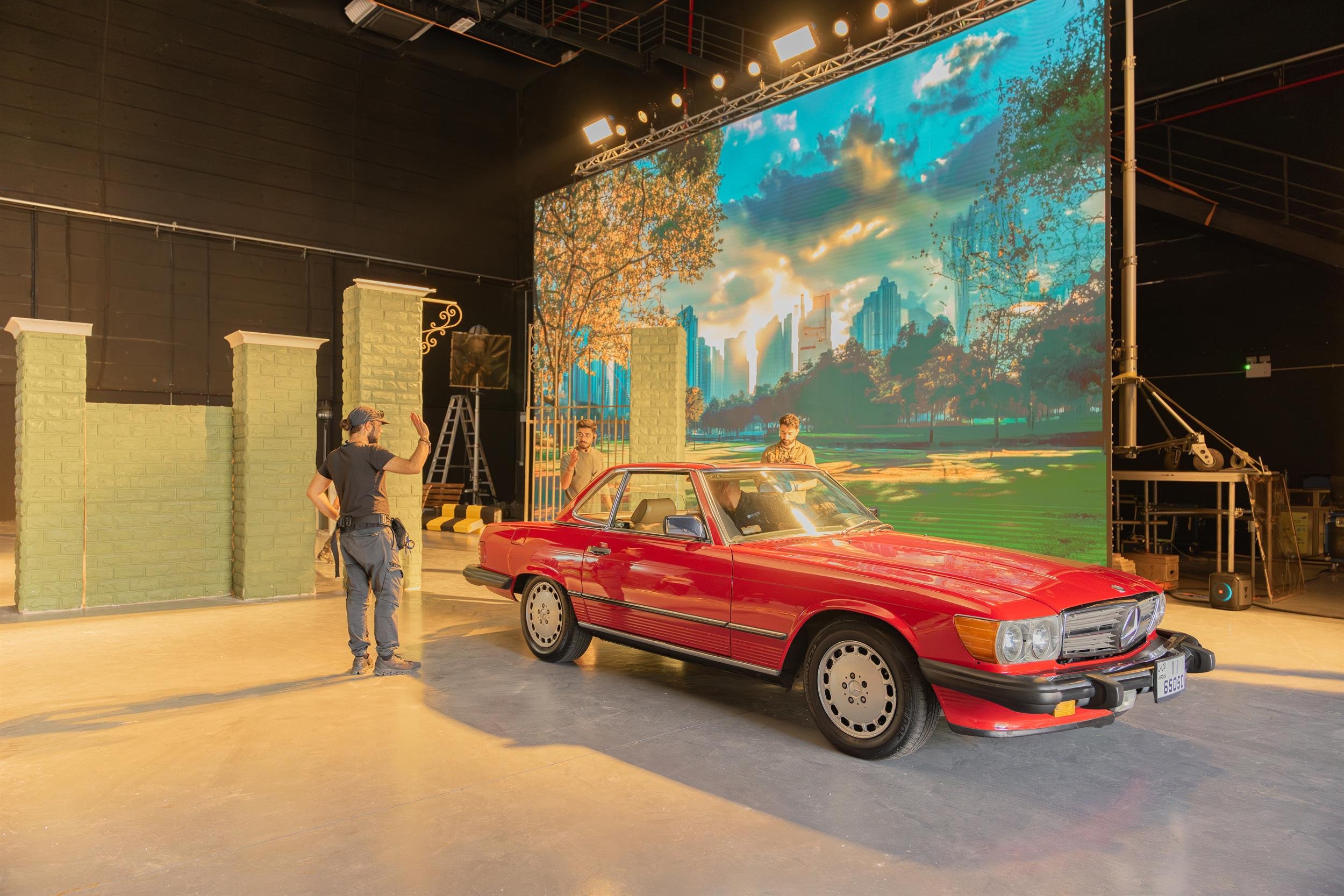Ahlan Simsim unit still: Crew members prepare a red vintage car in front of a large LED screen showing a city park.