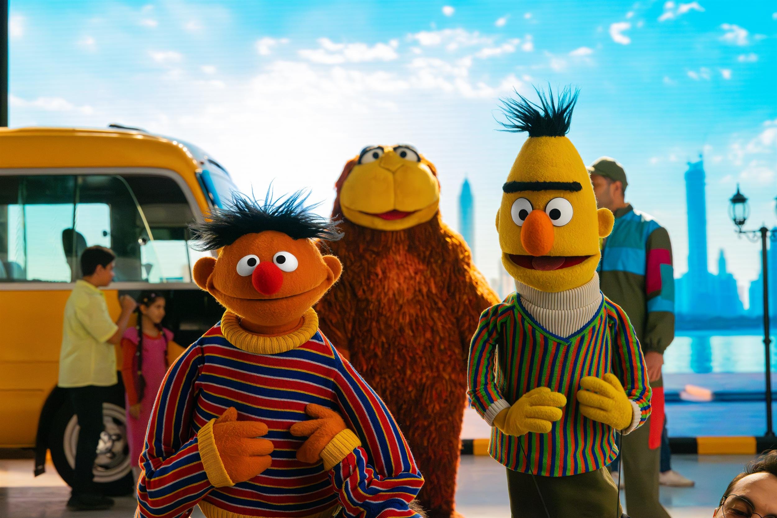Ahlan Simsim key art: Ernie, Bert and an orange bird muppet by the yellow bus on the city set.