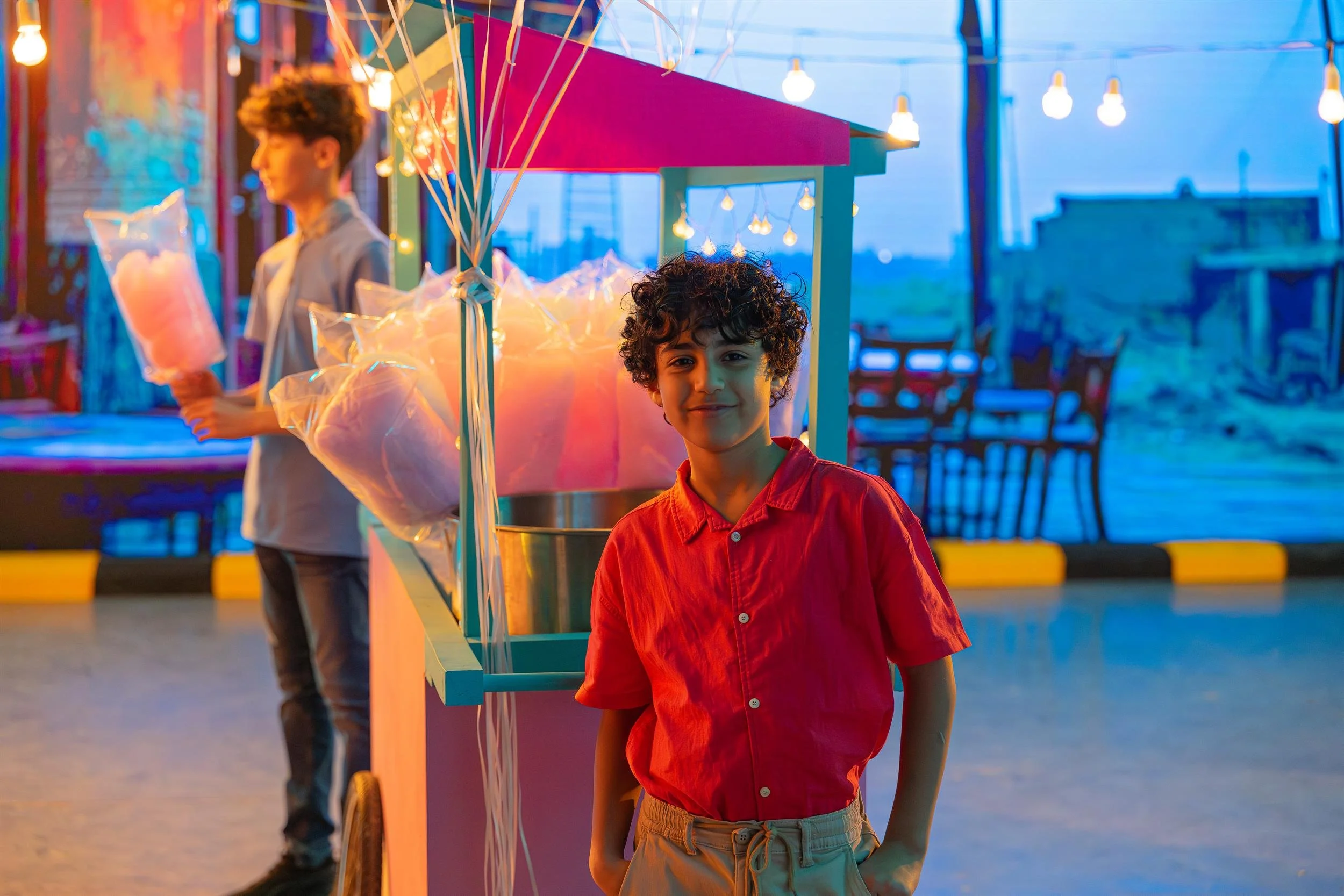 Ahlan Simsim unit still: a smiling boy stands in front of a colorful cotton candy cart with string lights.