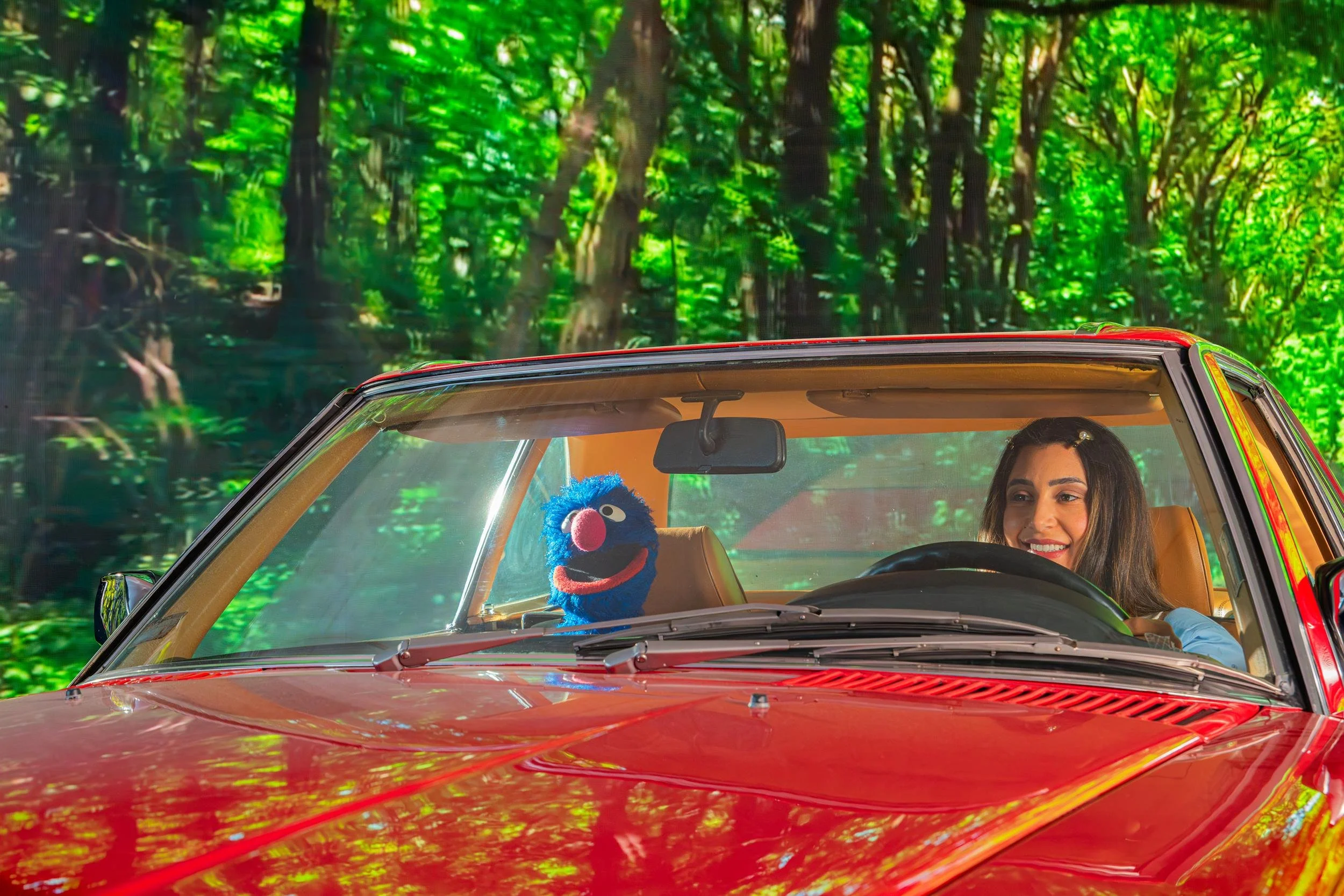 Ahlan Simsim key art: A smiling woman drives a red car with a blue Muppet in the back seat against a forest backdrop.