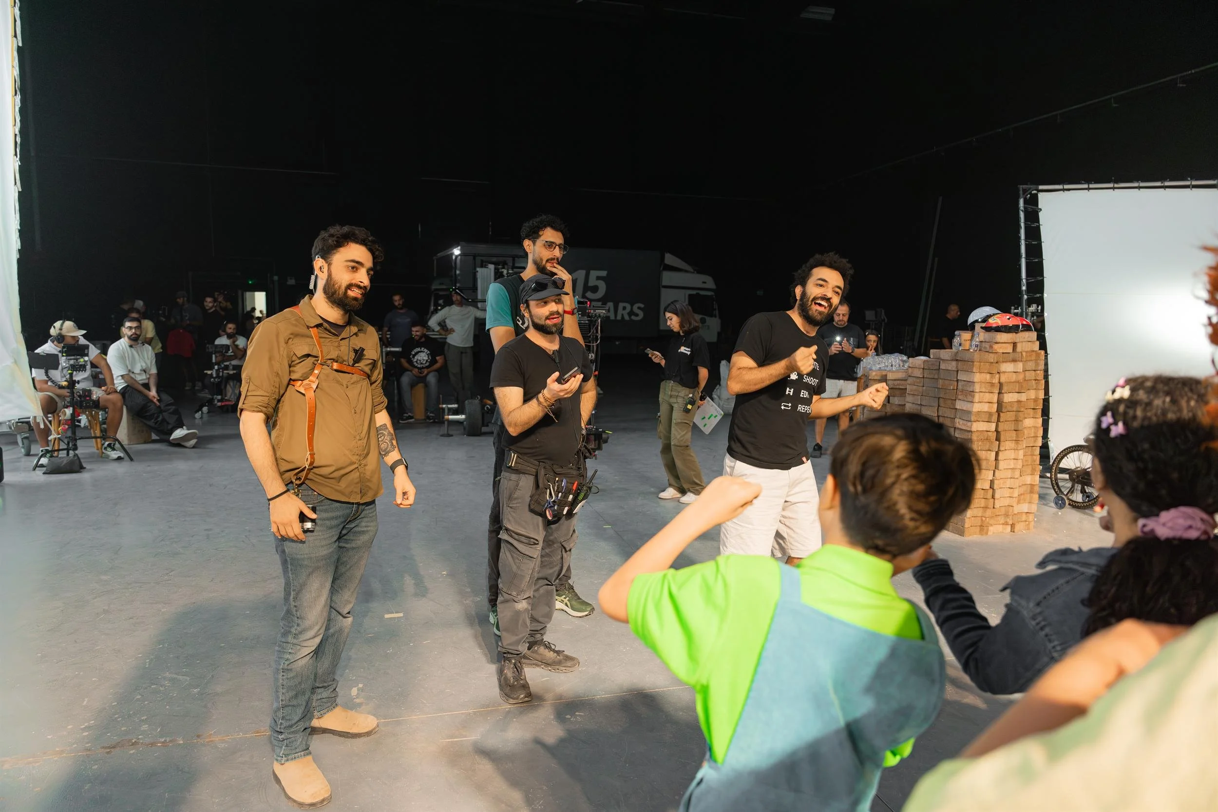 Ahlan Simsim unit still: Crew members talk with two children beside a tall brick-wall prop on a studio floor.