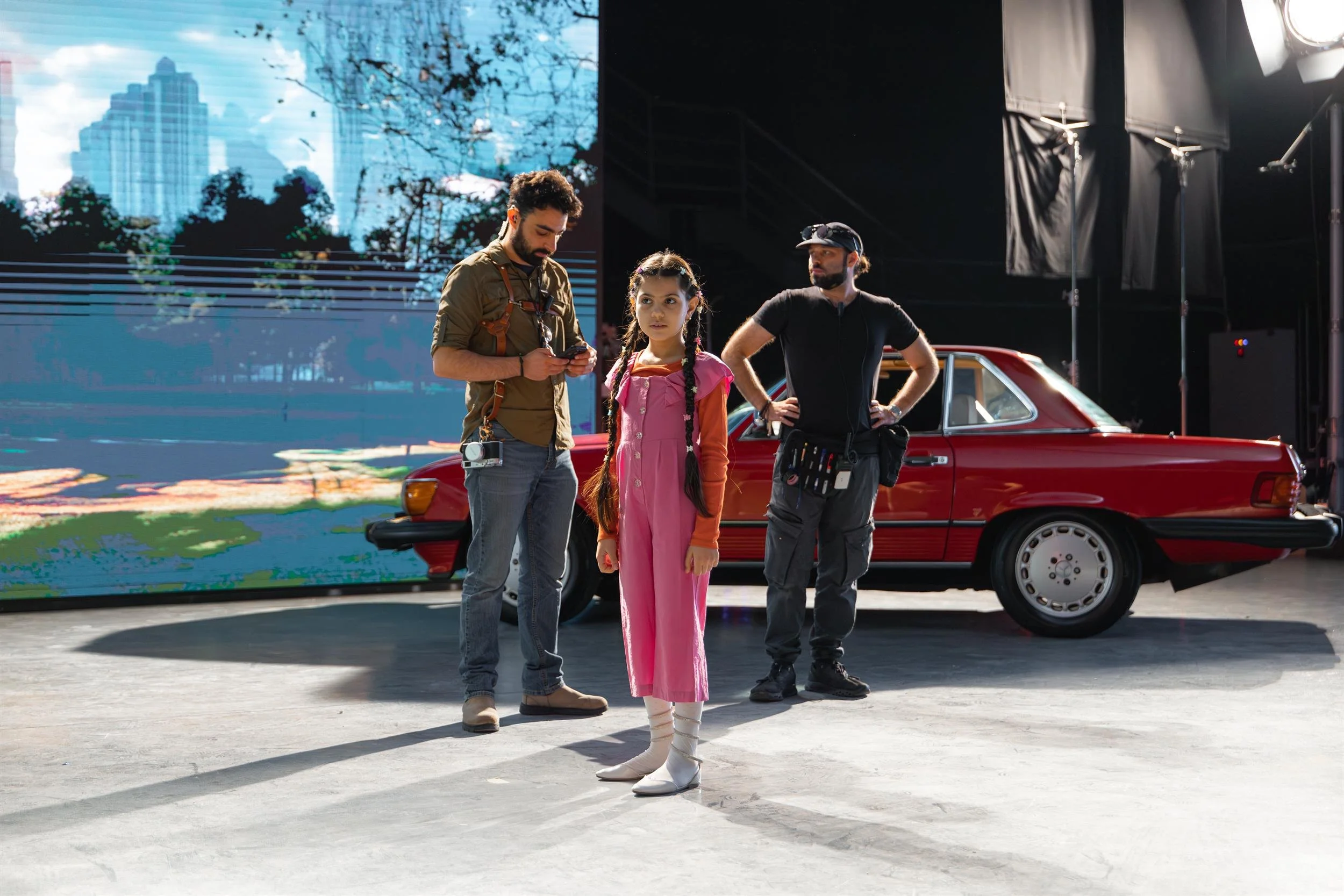 Ahlan Simsim unit still: A young girl stands between two crew members in front of a red car and a digital backdrop.