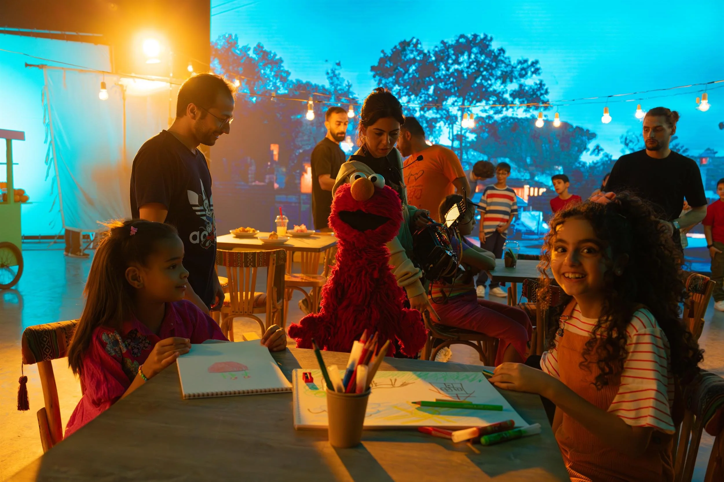 Ahlan Simsim unit still: Elmo and crew stand with two girls drawing at a table on a night-market set.