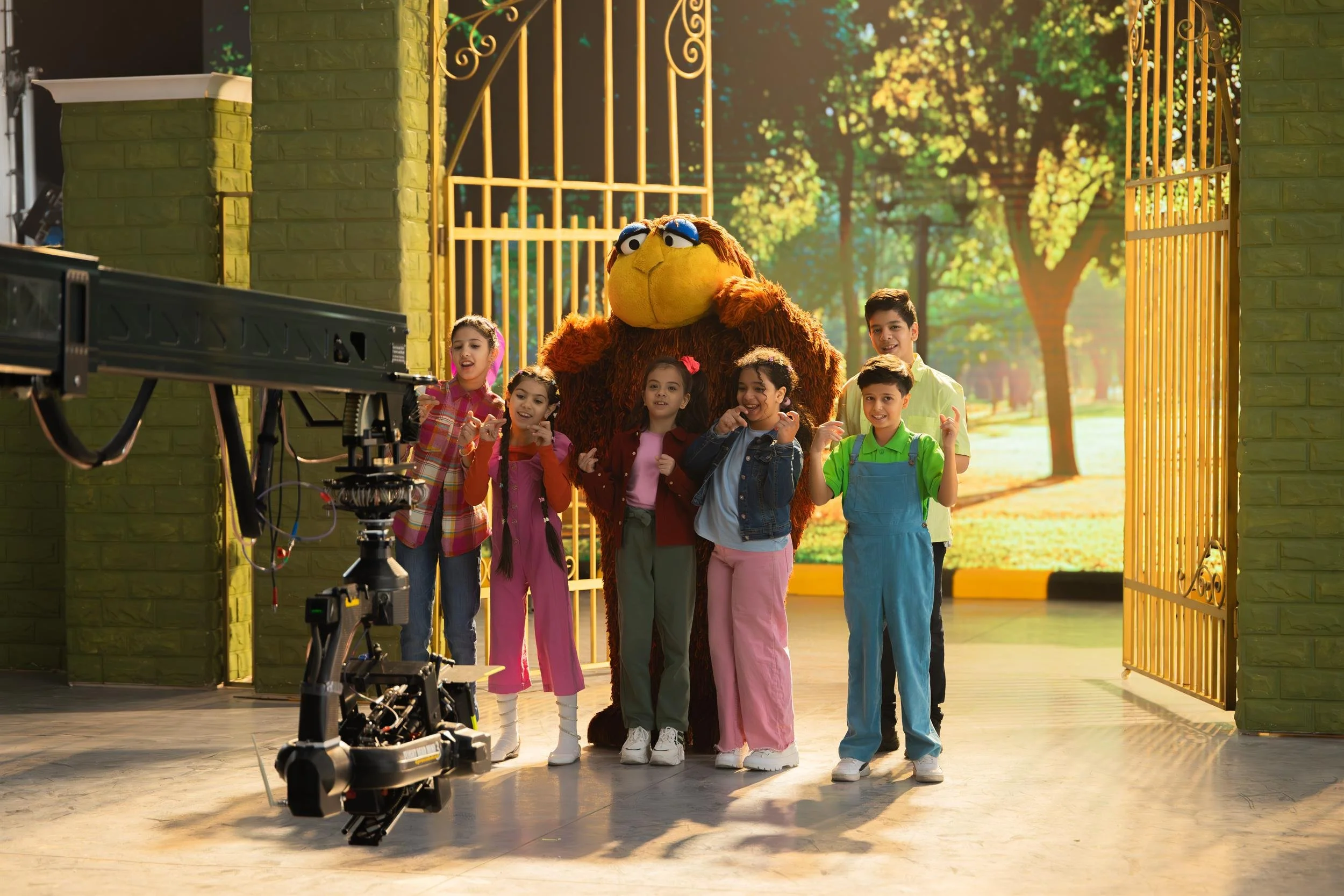 Ahlan Simsim unit still: Children and a large brown Muppet pose on a park set in front of a camera crane.