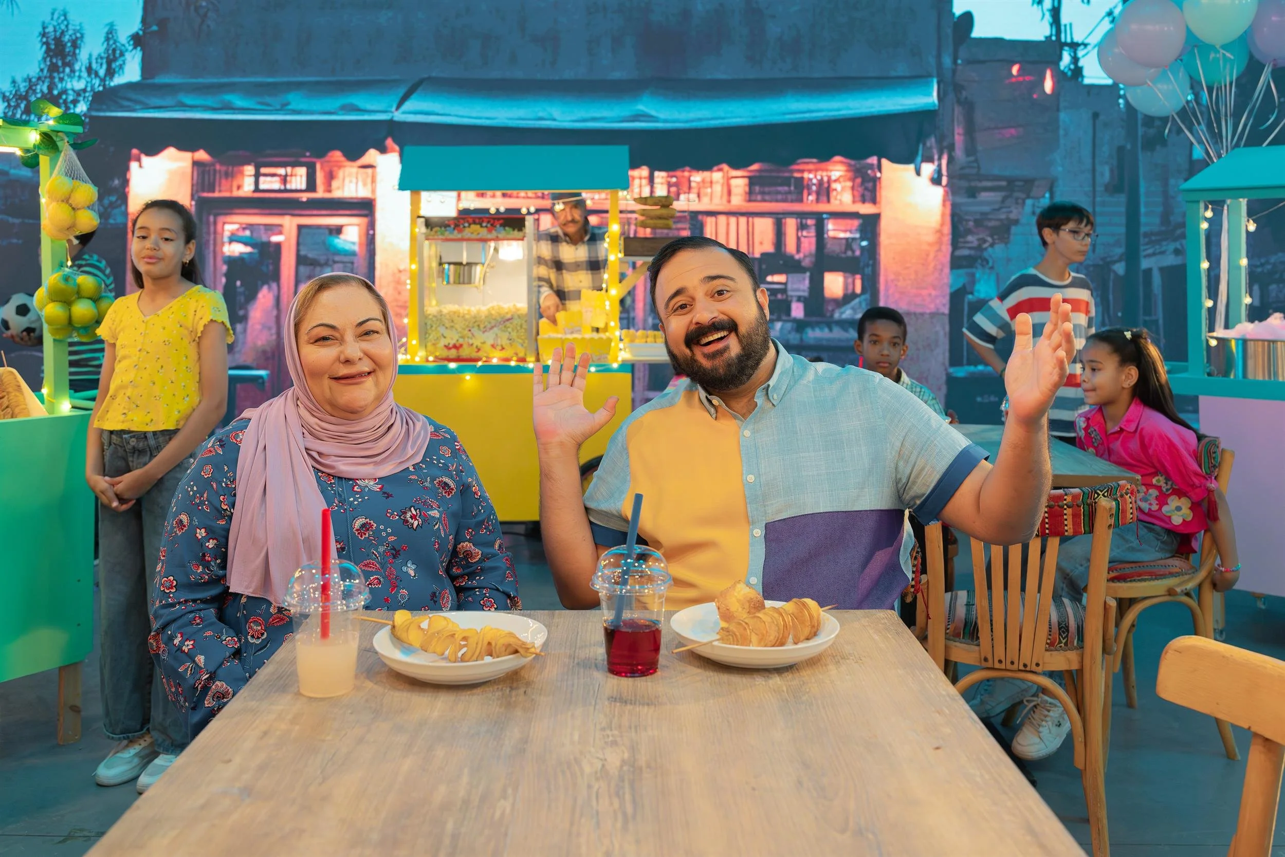 Ahlan Simsim key art: A man and a woman sit at a wooden table with snacks in a festive outdoor market at dusk.