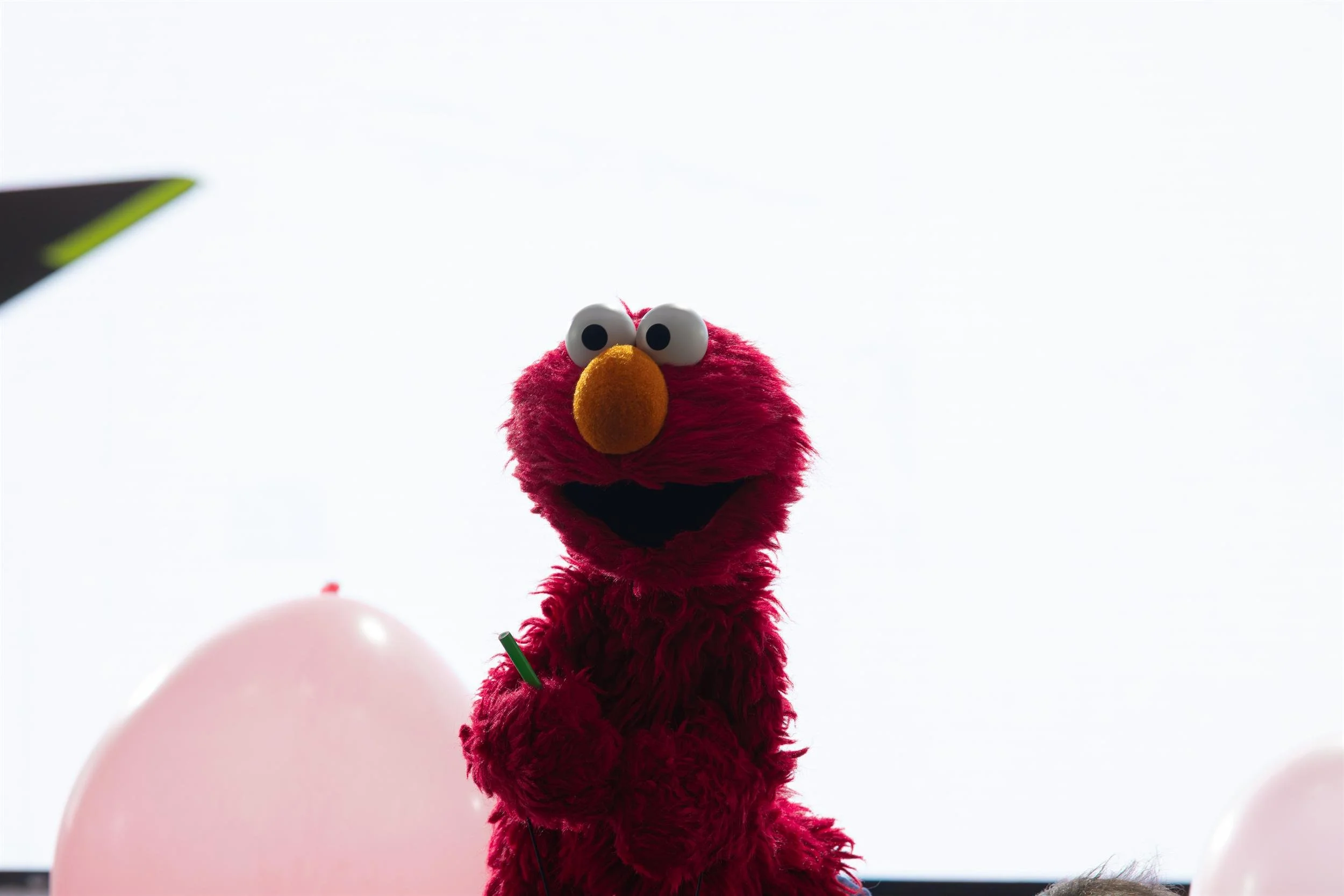 Ahlan Simsim unit still: Elmo holds a green stick against a bright white backdrop, with a pink balloon in the foreground.