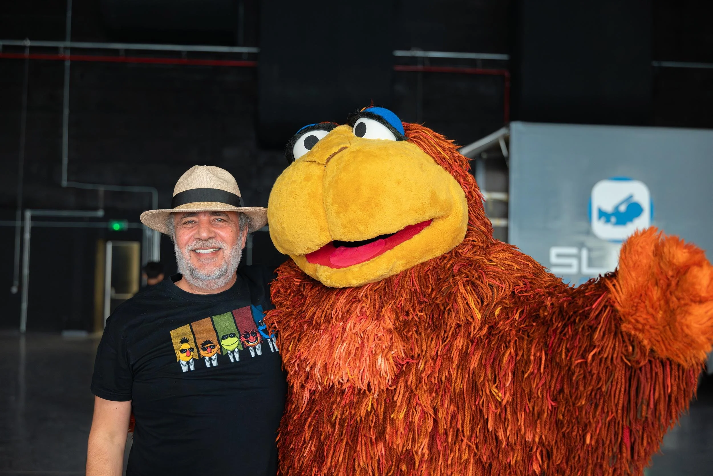 Ahlan Simsim unit still: a man in a fedora poses beside a large shaggy brown Muppet waving at the camera.