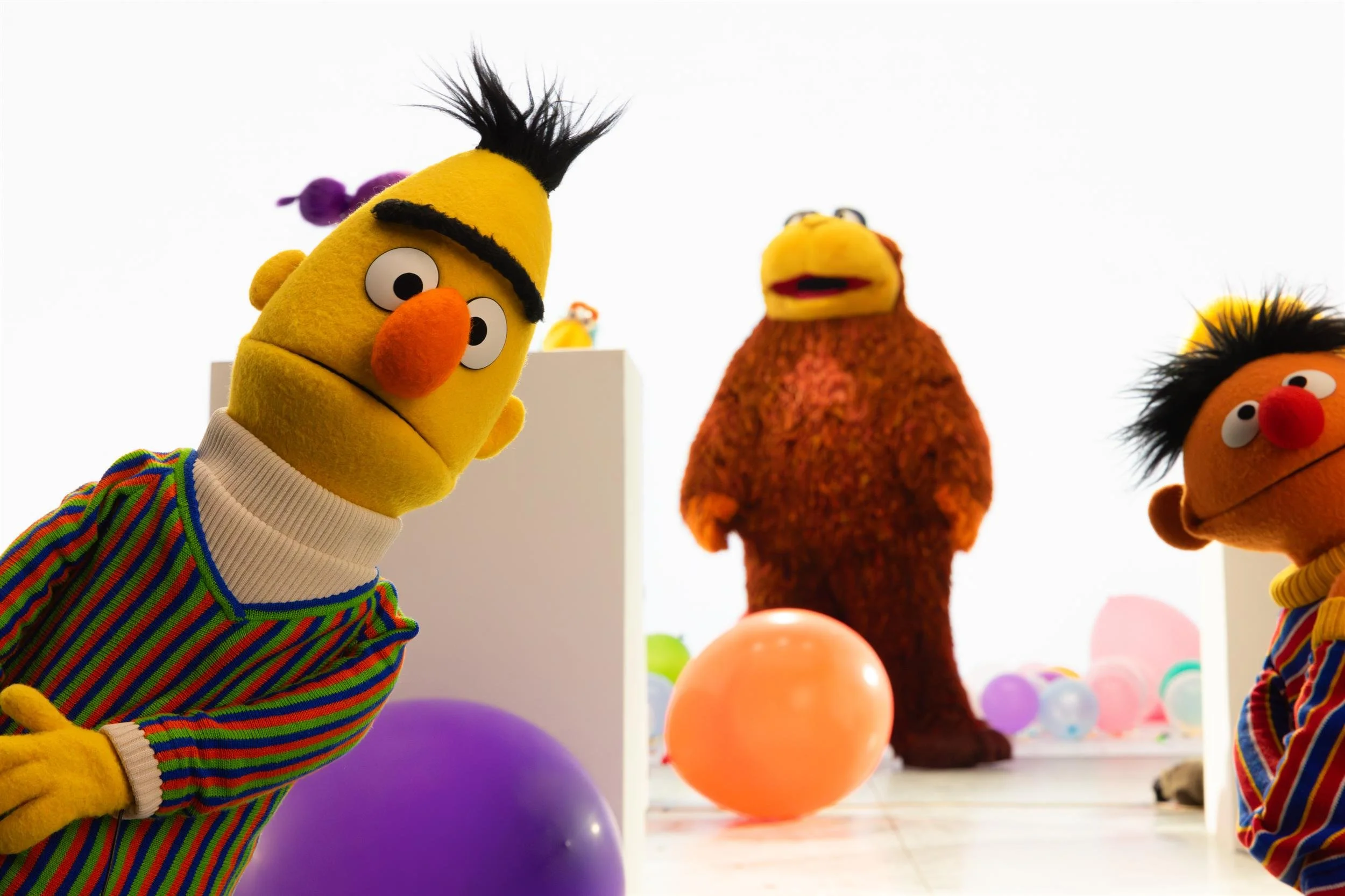 Ahlan Simsim unit still: Bert and Ernie lean into the frame on a bright white set with a large brown Muppet and balloons.