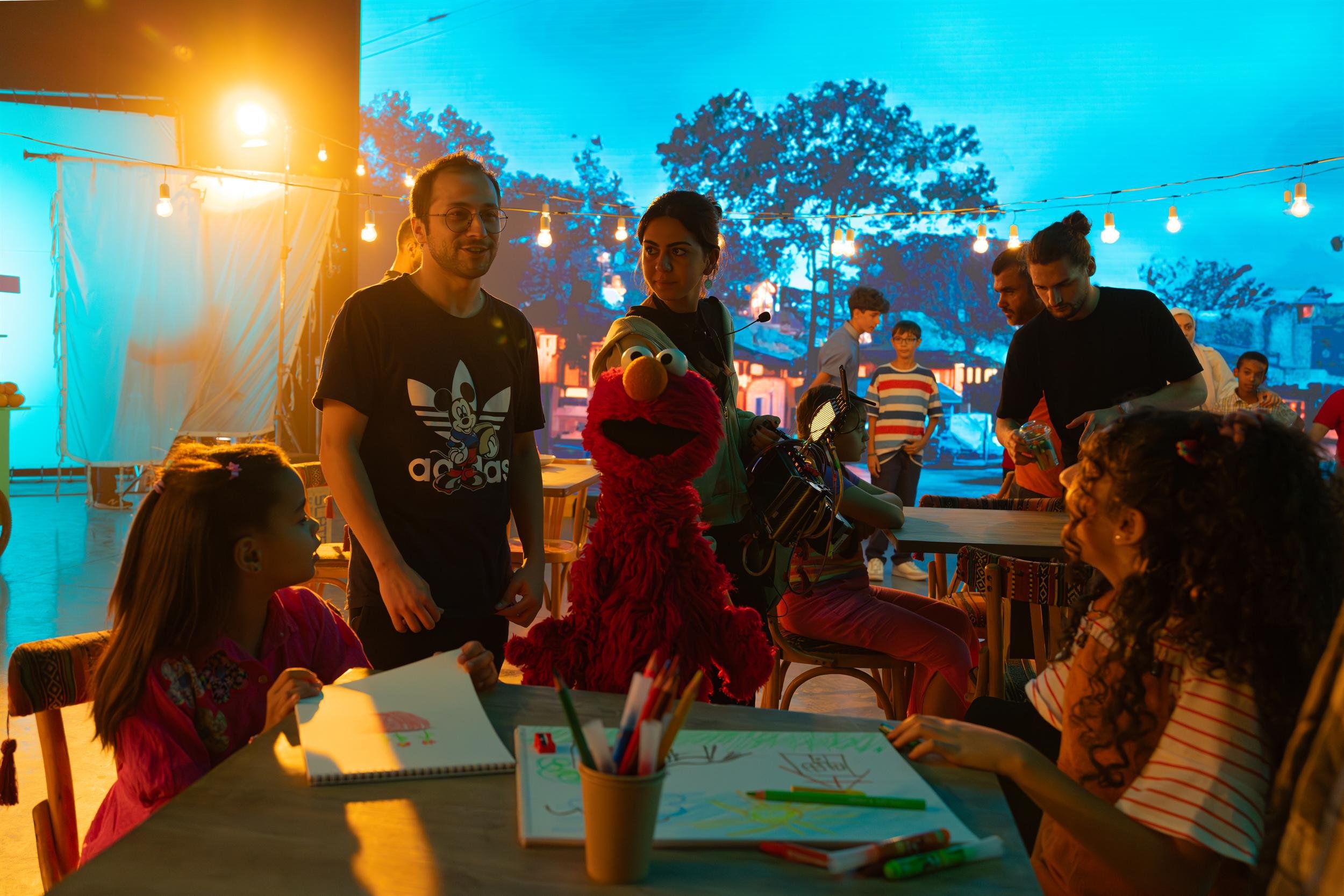 Ahlan Simsim unit still: crew and children film a scene with Elmo at a table under string lights.