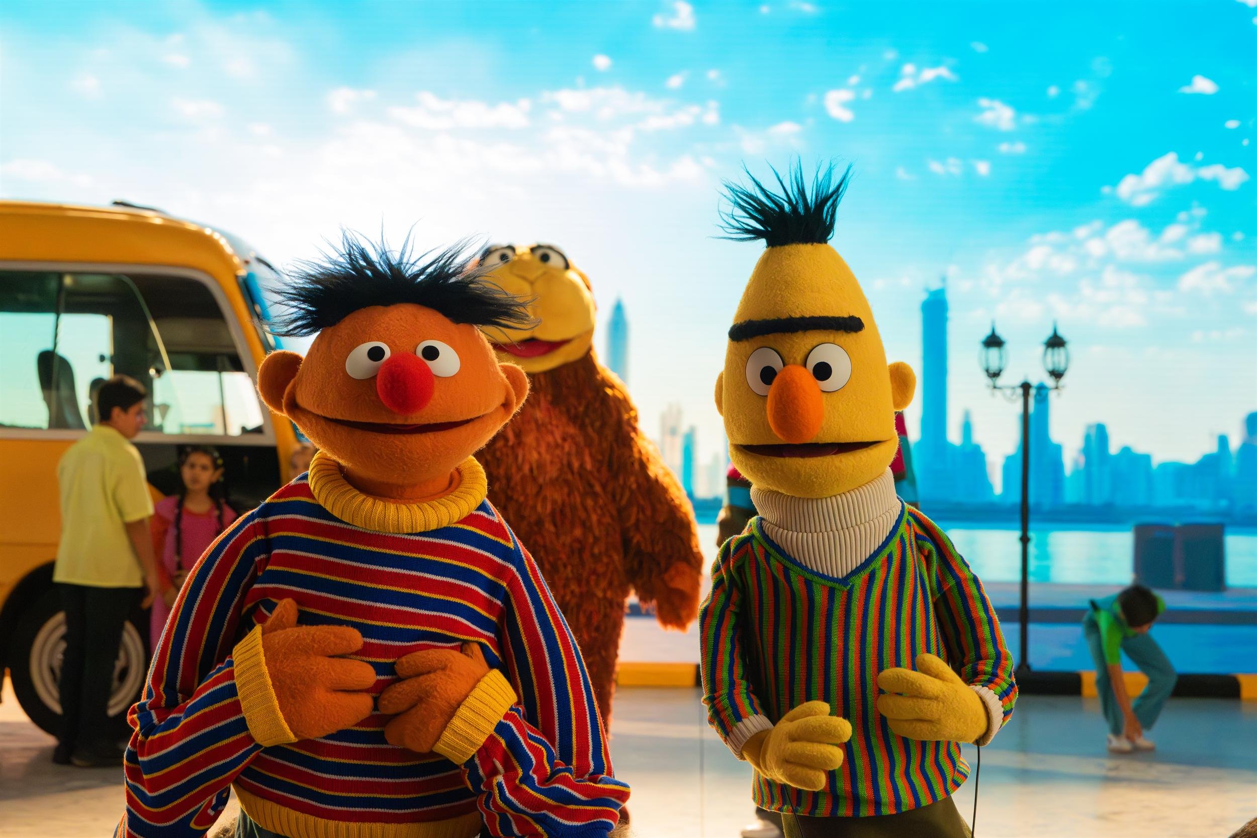 Ahlan Simsim unit still: Ernie, Bert, and a large brown Muppet pose with children near a yellow bus against a city skyline.