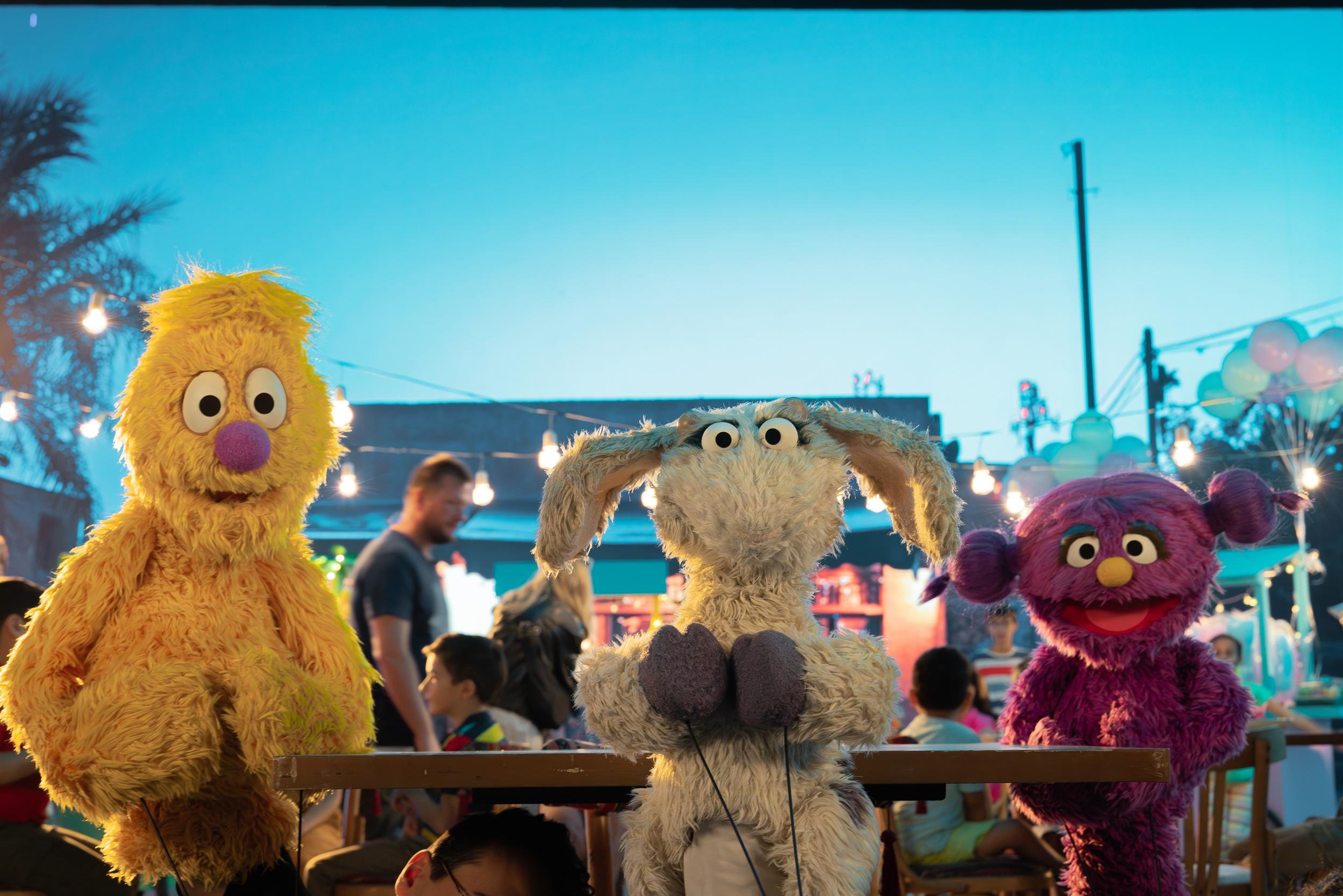 Ahlan Simsim unit still: a yellow Muppet, a goat Muppet, and a purple Muppet sit at a wooden table on a night-market set.
