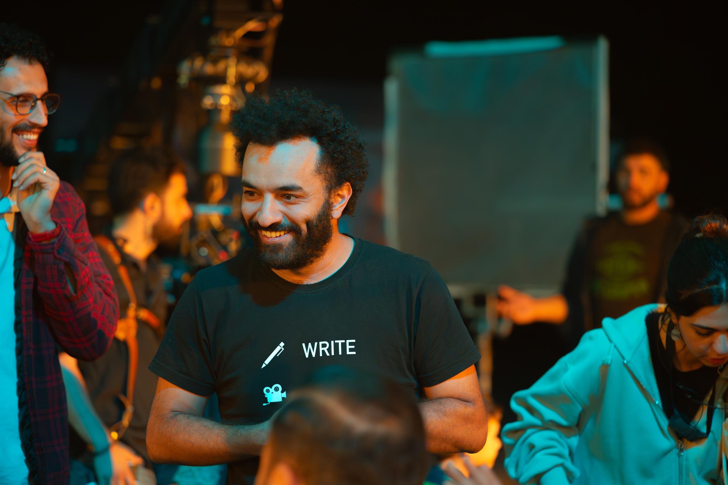 Ahlan Simsim unit still: a smiling crew member in a black t-shirt stands among other crew on a dim set.