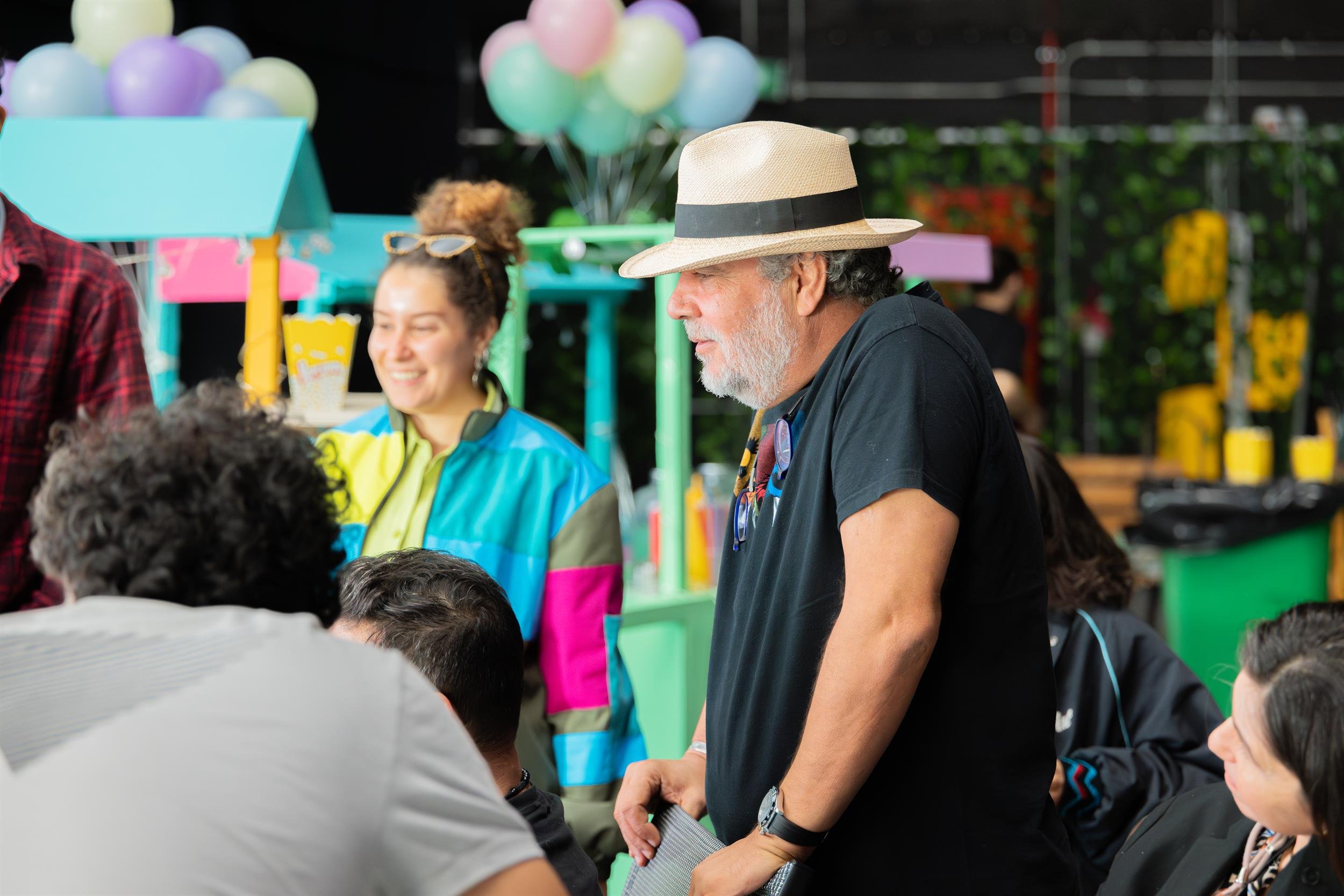 Ahlan Simsim unit still: crew members talk on a colorful market set decorated with pastel balloons.