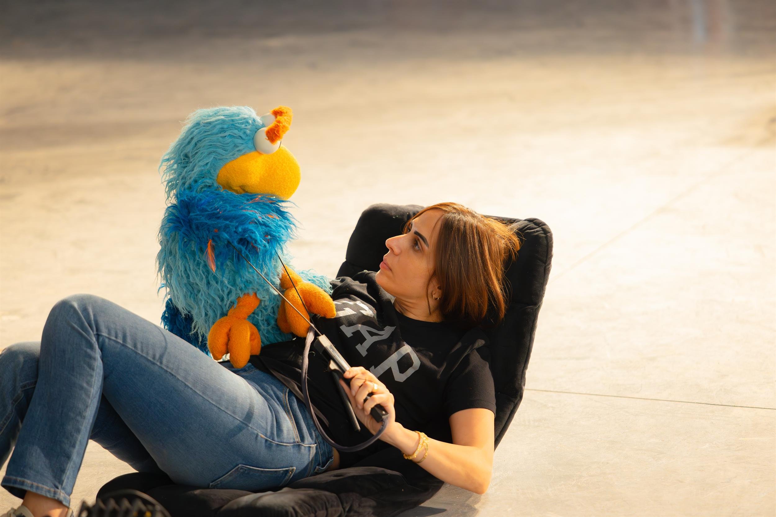 Ahlan Simsim unit still: a puppeteer reclines on a floor chair holding a blue bird Muppet.