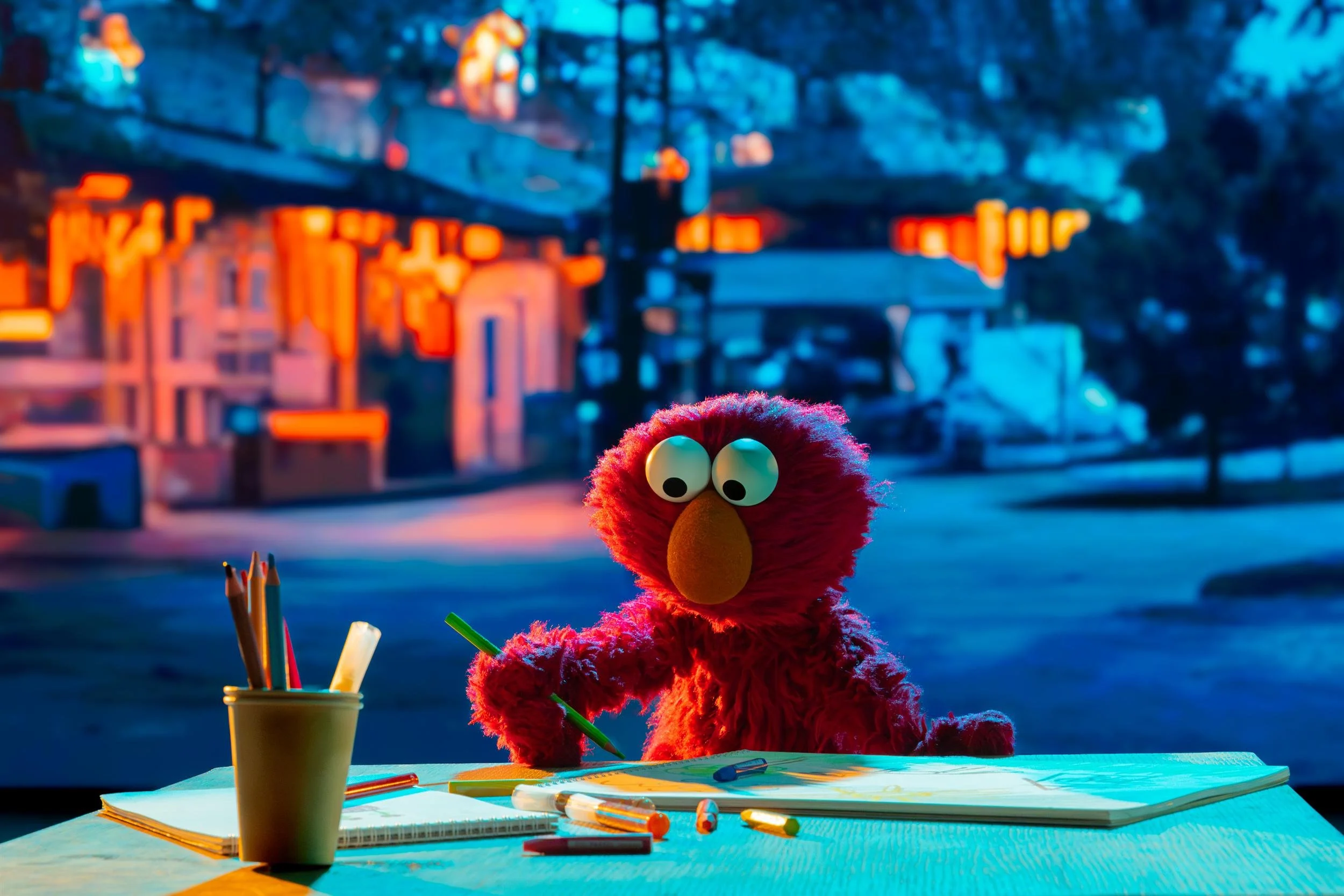 Ahlan Simsim key art: Elmo sits at a table and draws in a notebook against a nighttime city background.