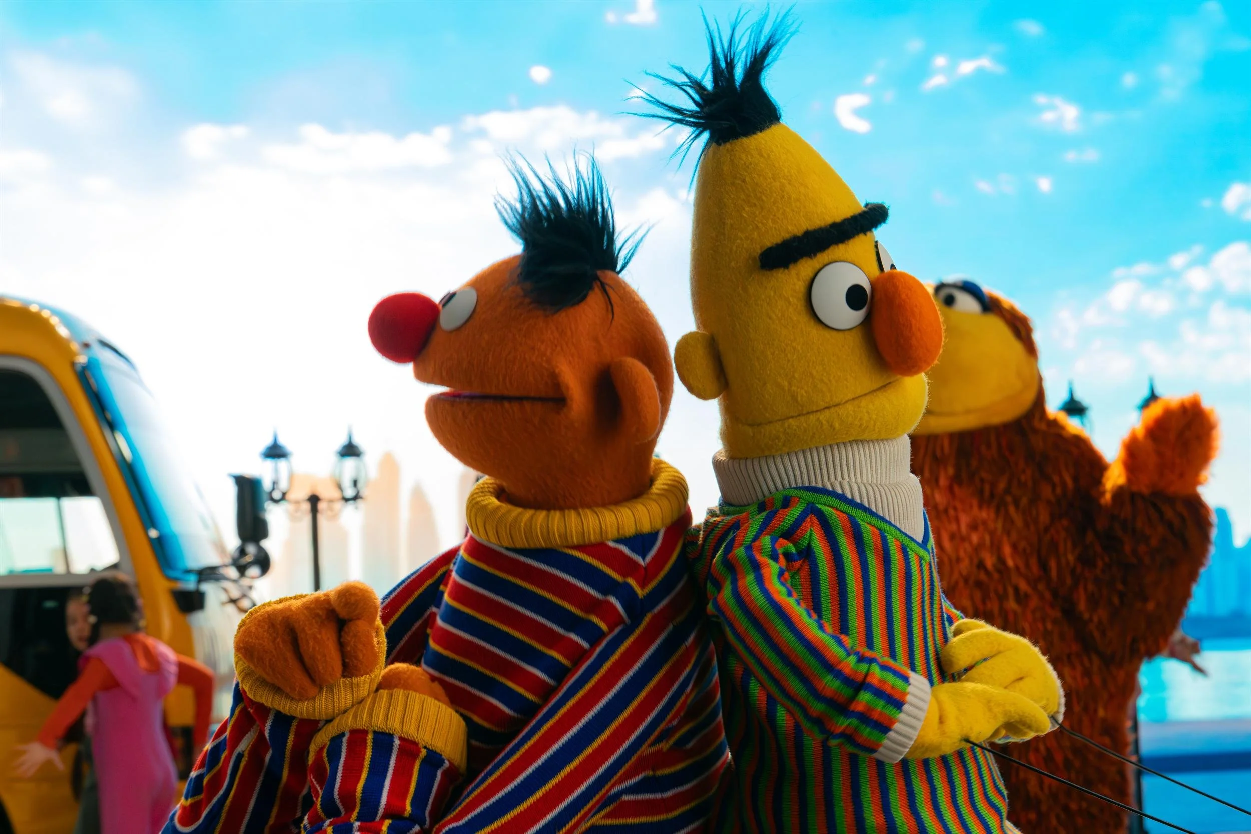 Ahlan Simsim unit still: Ernie and Bert stand back-to-back near a yellow bus, with a brown Muppet behind them.