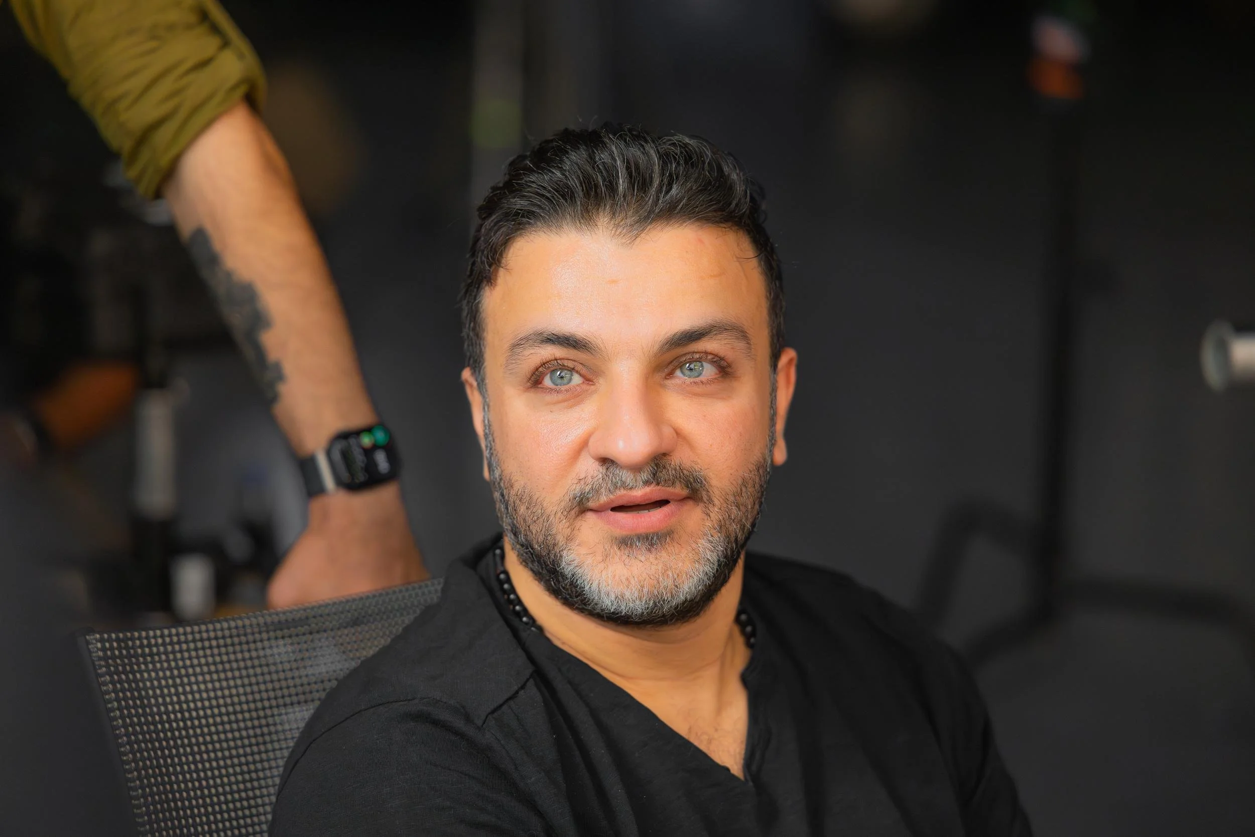 Ahlan Simsim unit still: A bearded man in a black shirt sits in a mesh chair on a studio set.