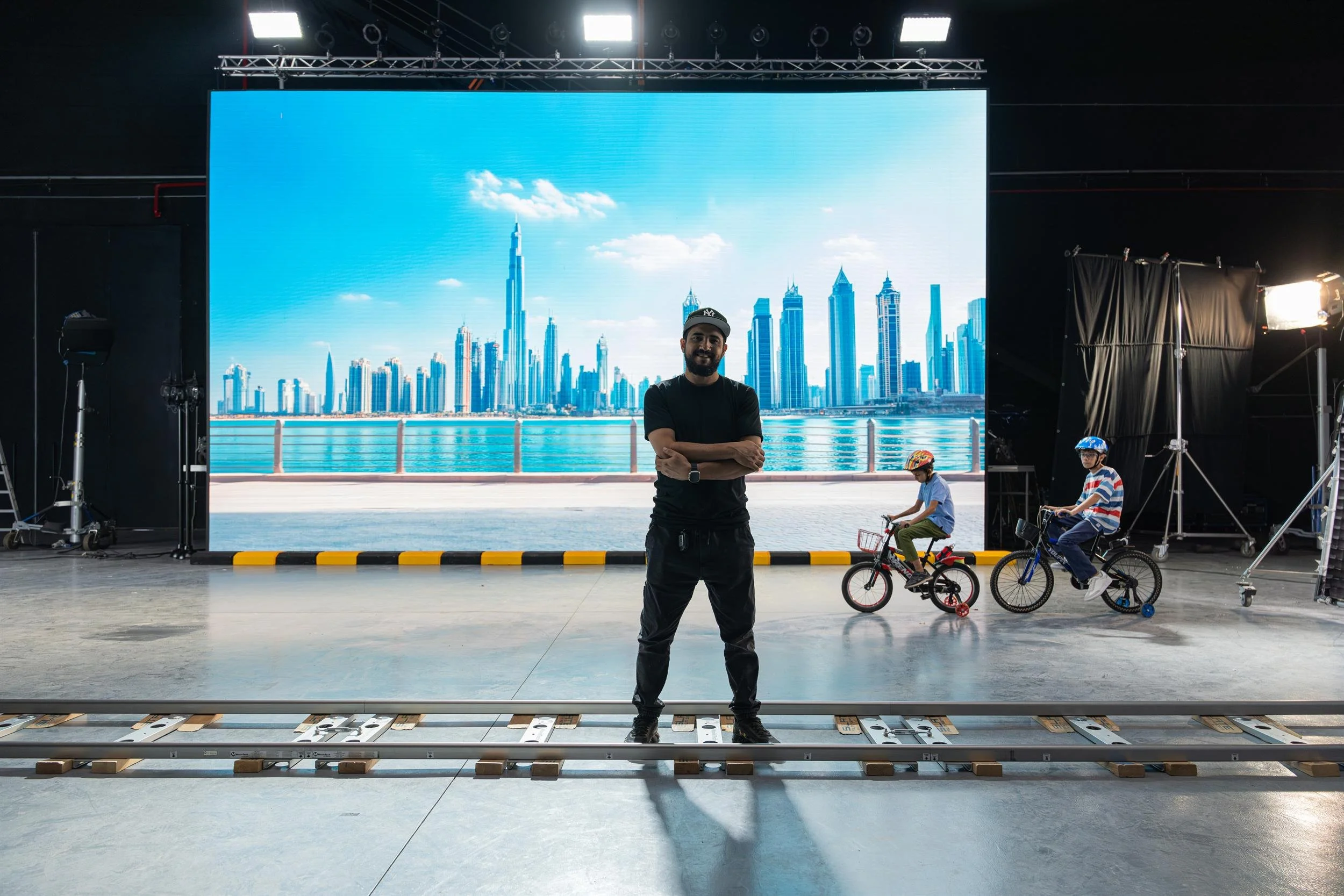 Ahlan Simsim unit still: a man stands on camera tracks before an LED city skyline, with two children on bicycles.