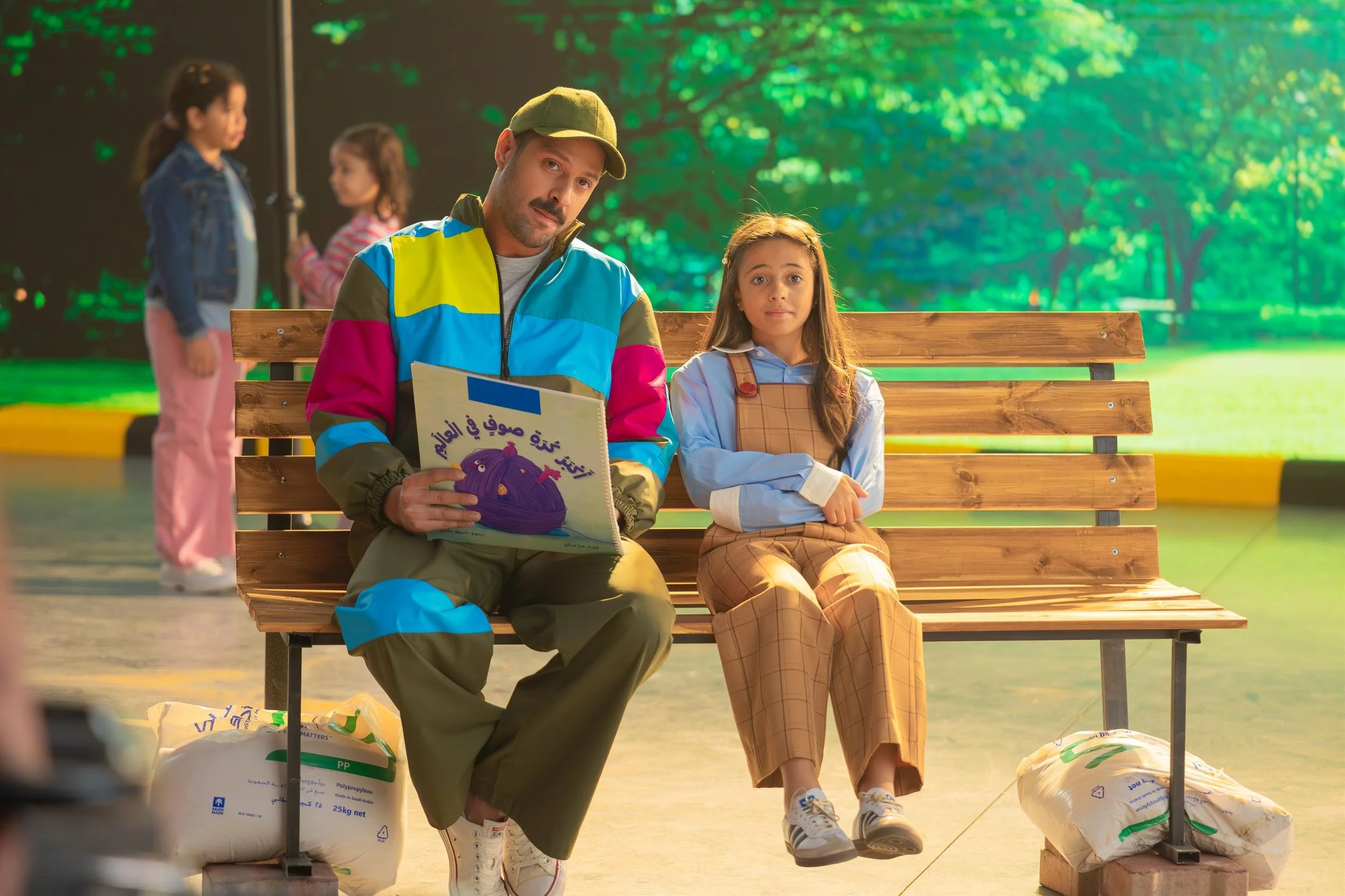 Ahlan Simsim unit still: A man in a colorful tracksuit and a girl sit on a wooden park bench, the man holding a children's book.