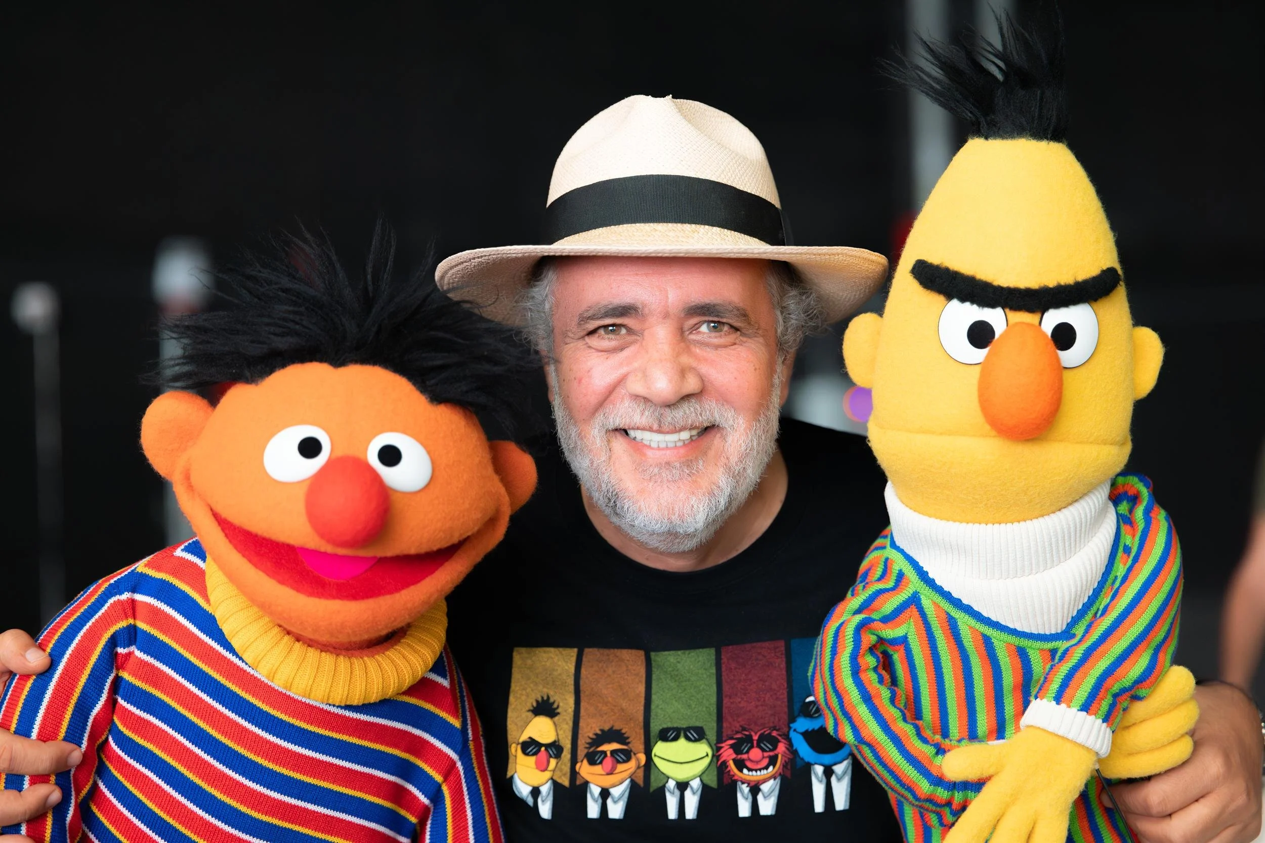 Ahlan Simsim unit still: a man in a fedora poses between Ernie and Bert against a dark studio background.