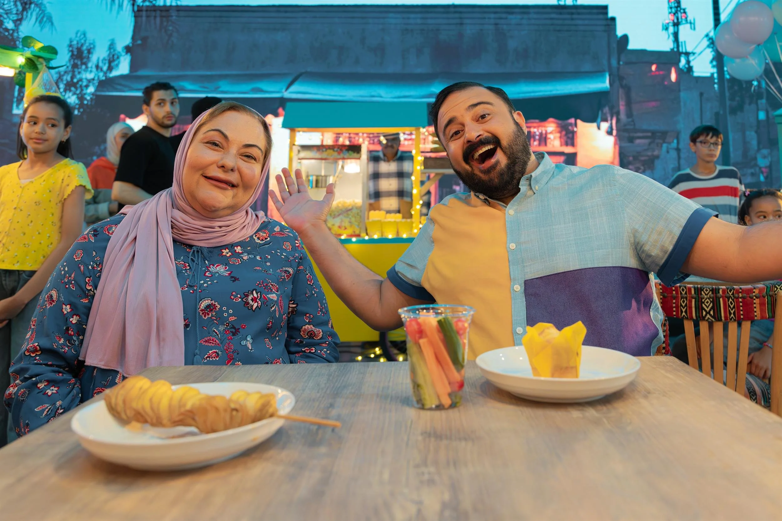 Ahlan Simsim unit still: a man and a woman sit at a table with snacks in front of a brightly lit food stall.